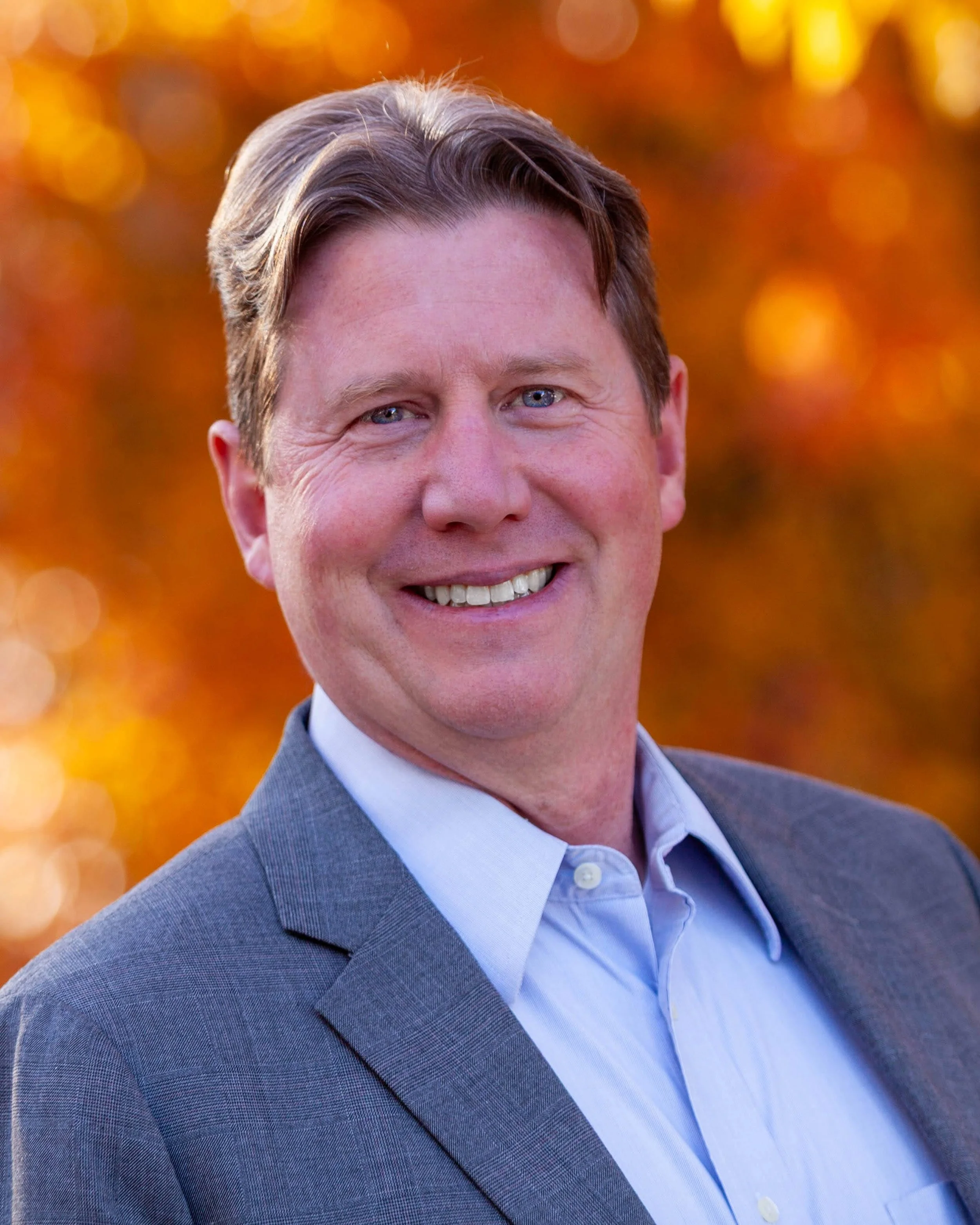A man in a gray suit smiling outdoors with a blurred background of orange and yellow autumn leaves.
