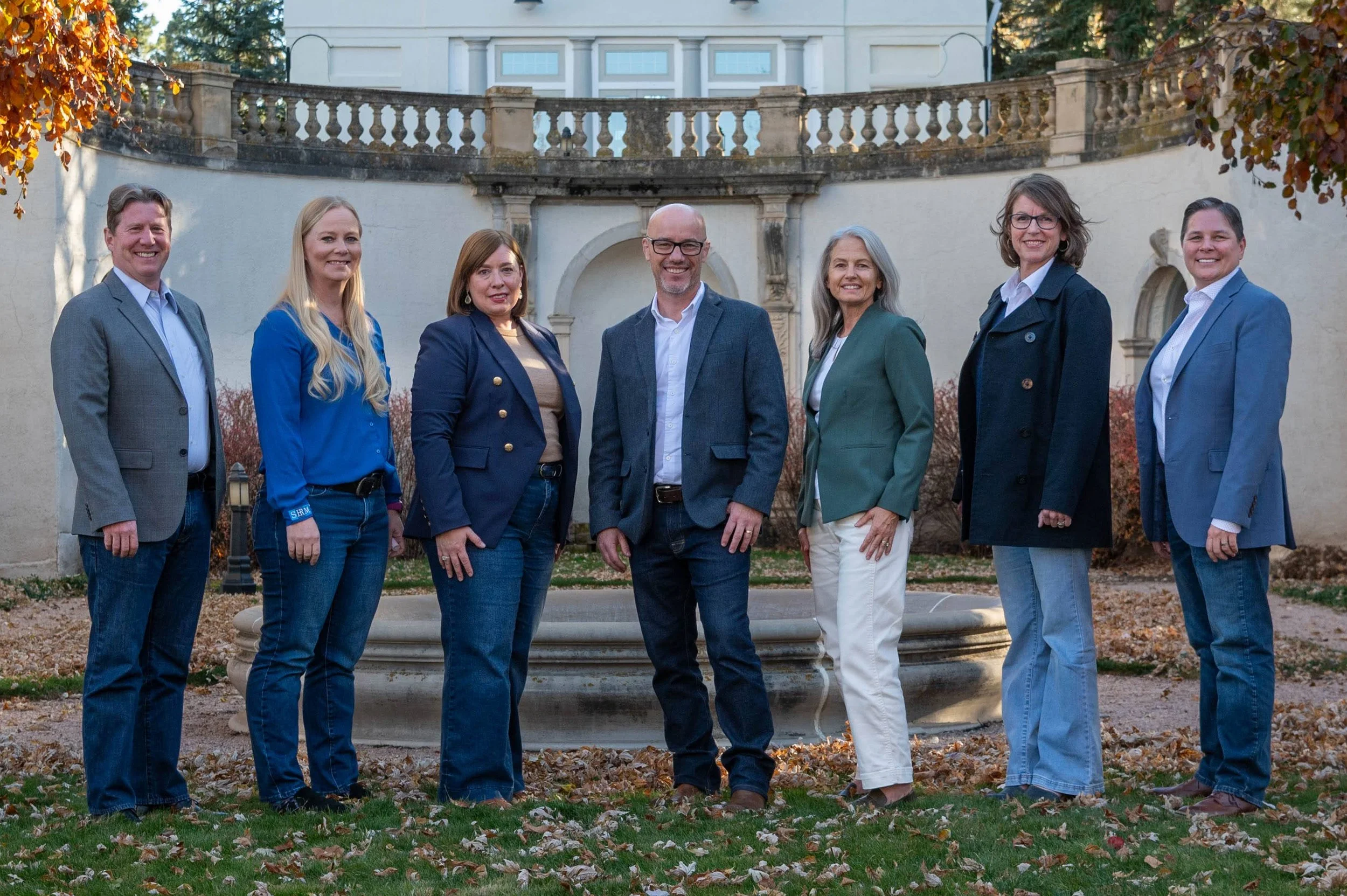 Group of eight people, five women and three men, standing outdoors in front of a fountain and a white building, dressed in business casual attire, smiling.