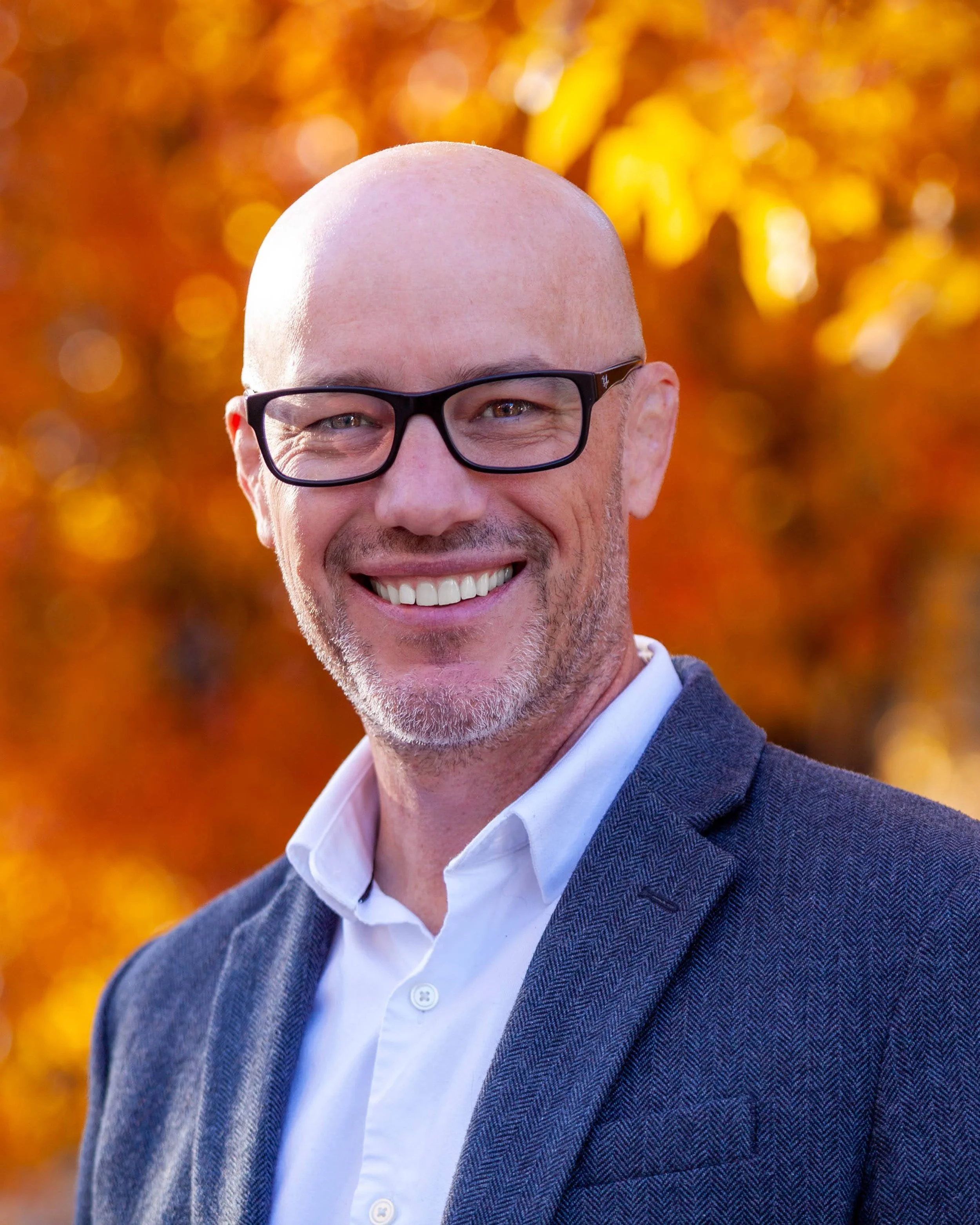 A smiling, bald man with glasses, wearing a white shirt and a blue blazer, standing outdoors with vibrant autumn foliage in the background.
