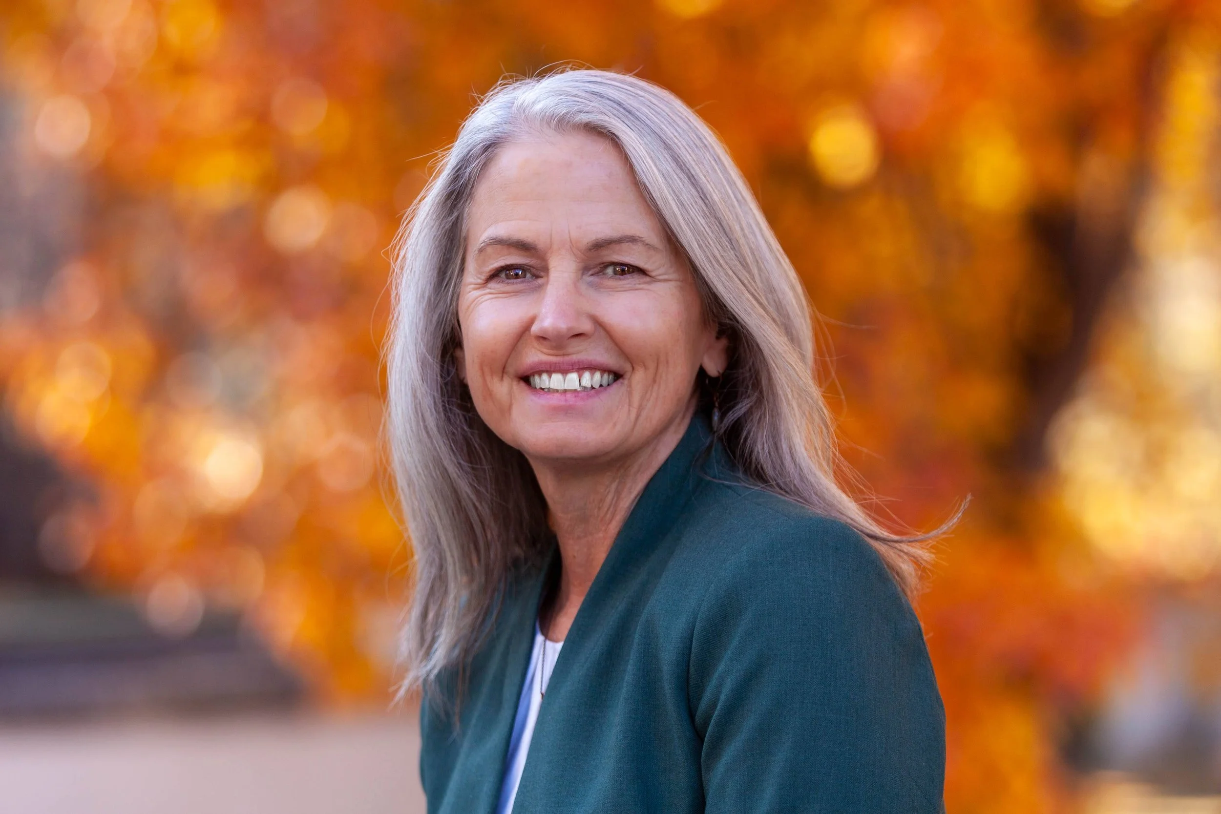 A smiling older woman with gray hair standing outdoors in front of orange autumn leaves.