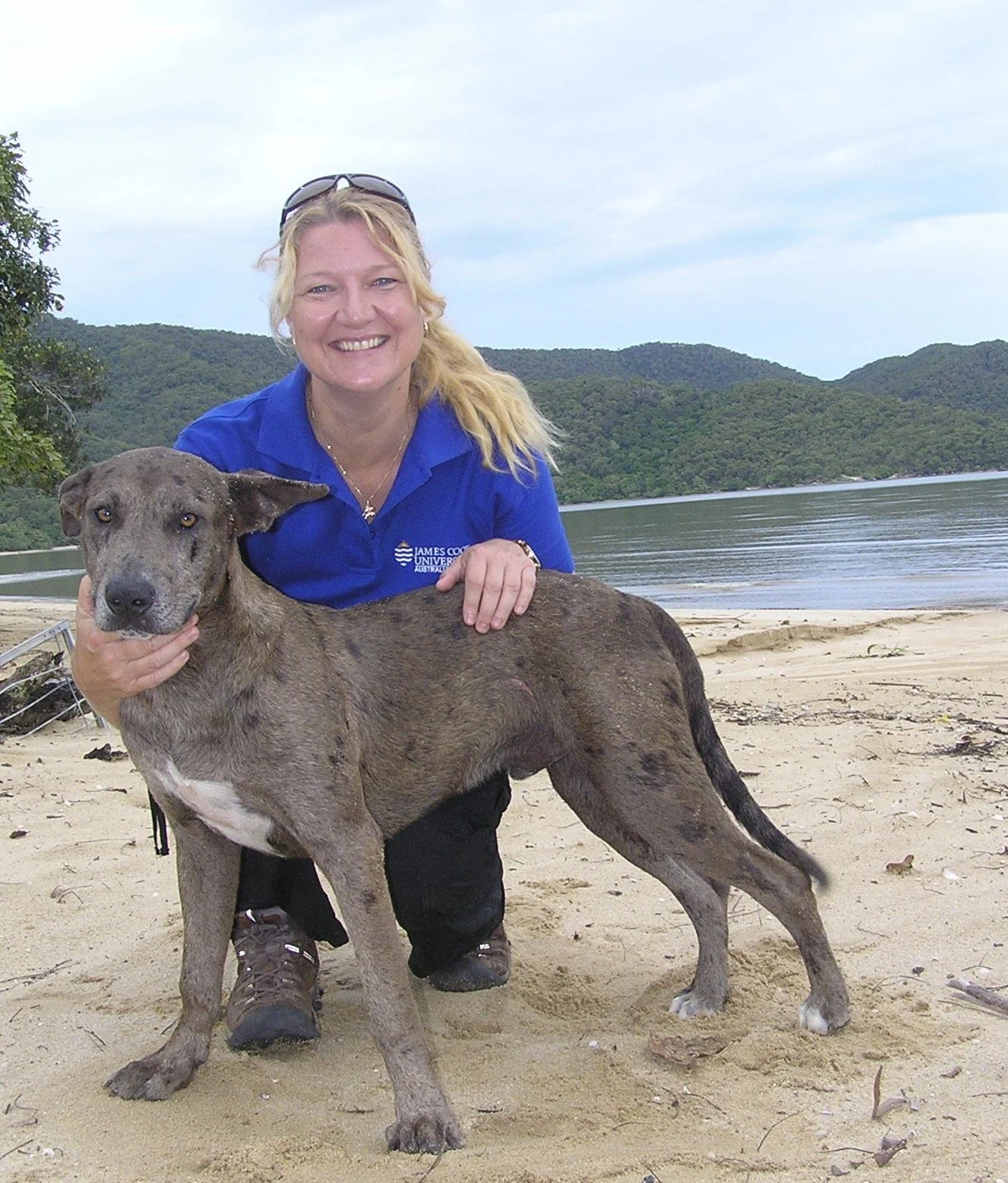 A woman in a blue shirt kneeling on a sandy beach with a large brindle-colored dog, both smiling, with a lake and green hills in the background.