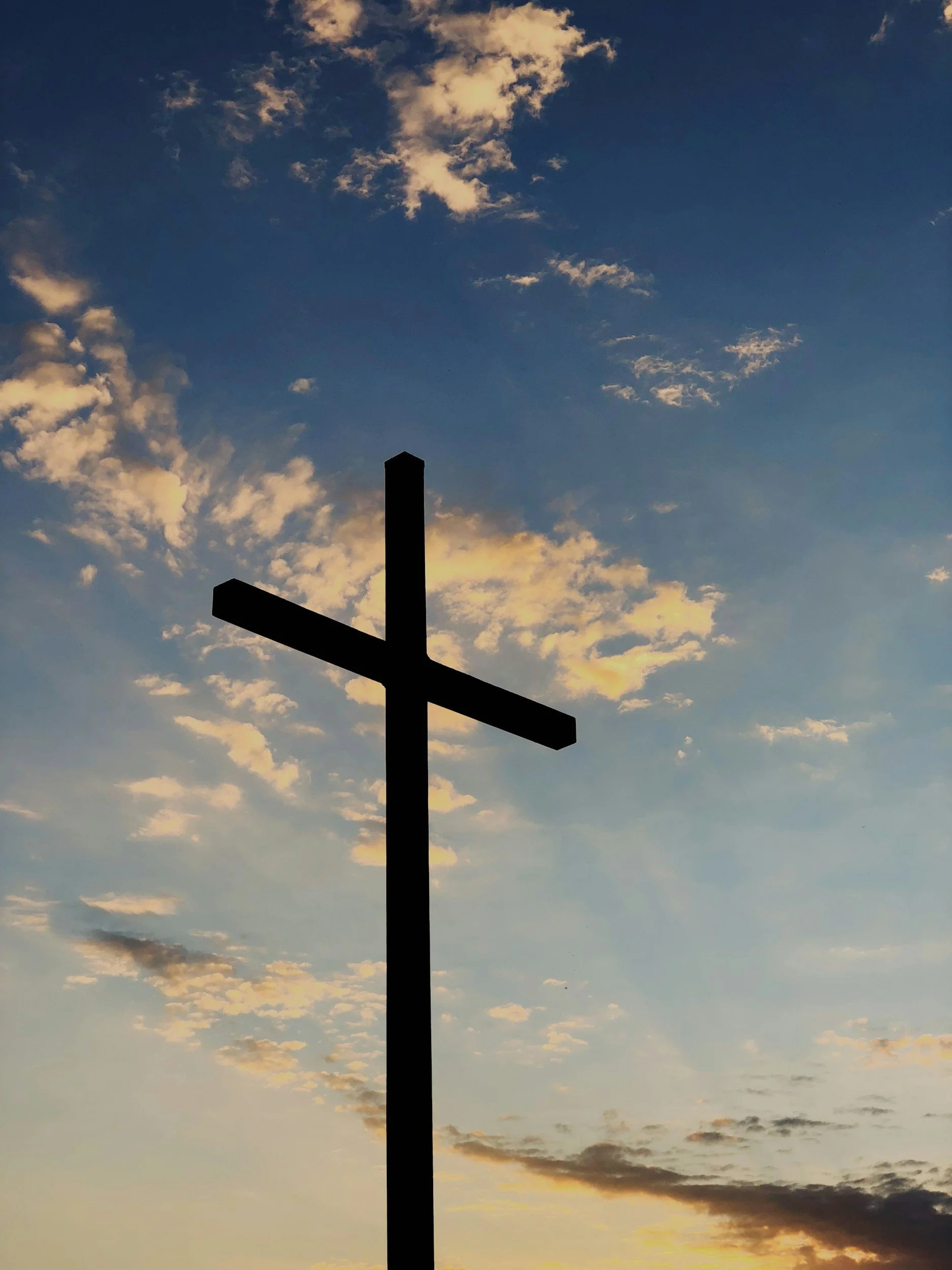 Silhouette of a Christian cross against a sky with scattered clouds during sunset or sunrise.
