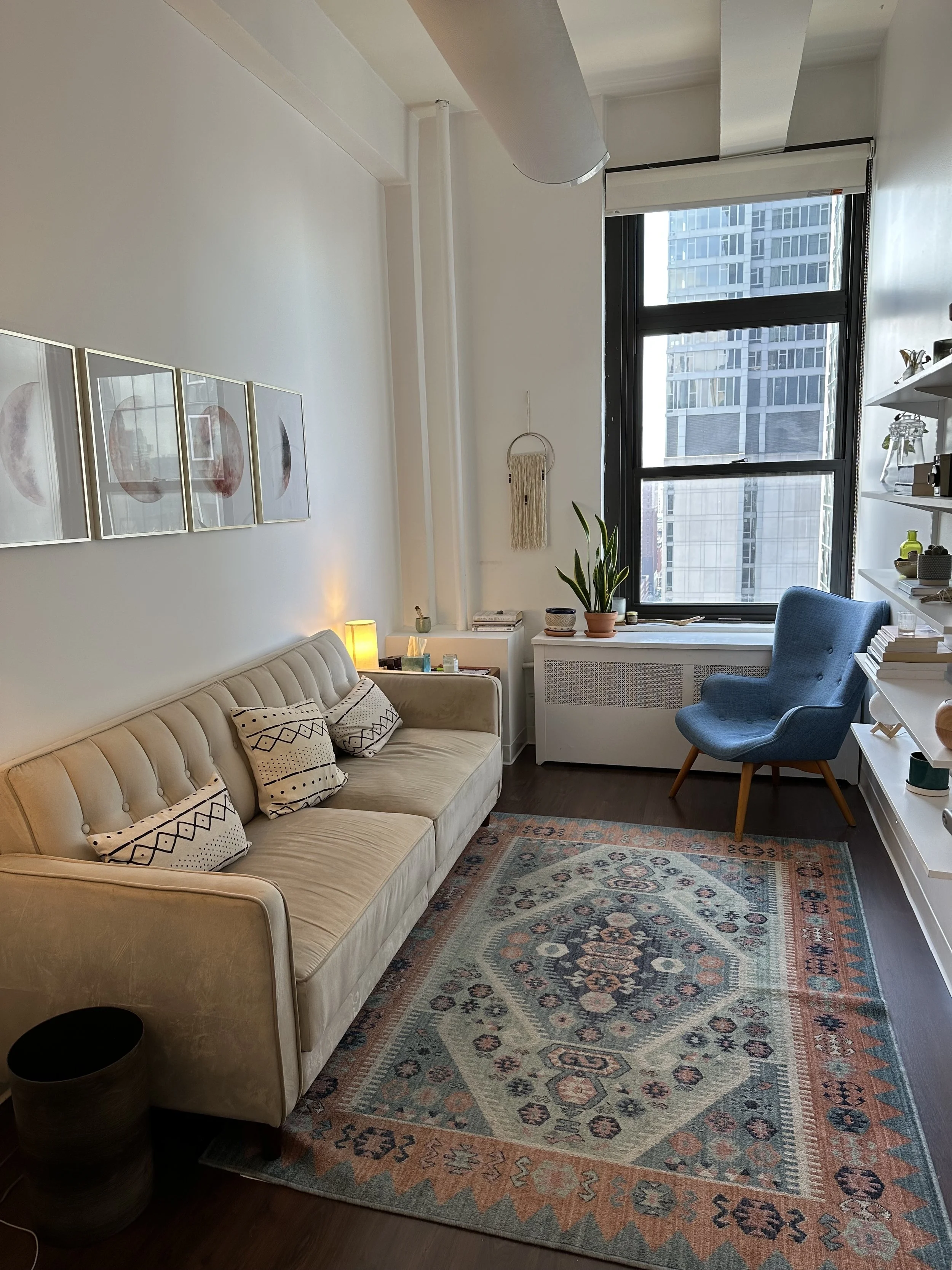 Living room with a beige tufted sofa, patterned pillows, a blue armchair near a large window, a white shelf with decor, a patterned rug, a wall hanging, and a small table with a lamp and plants.