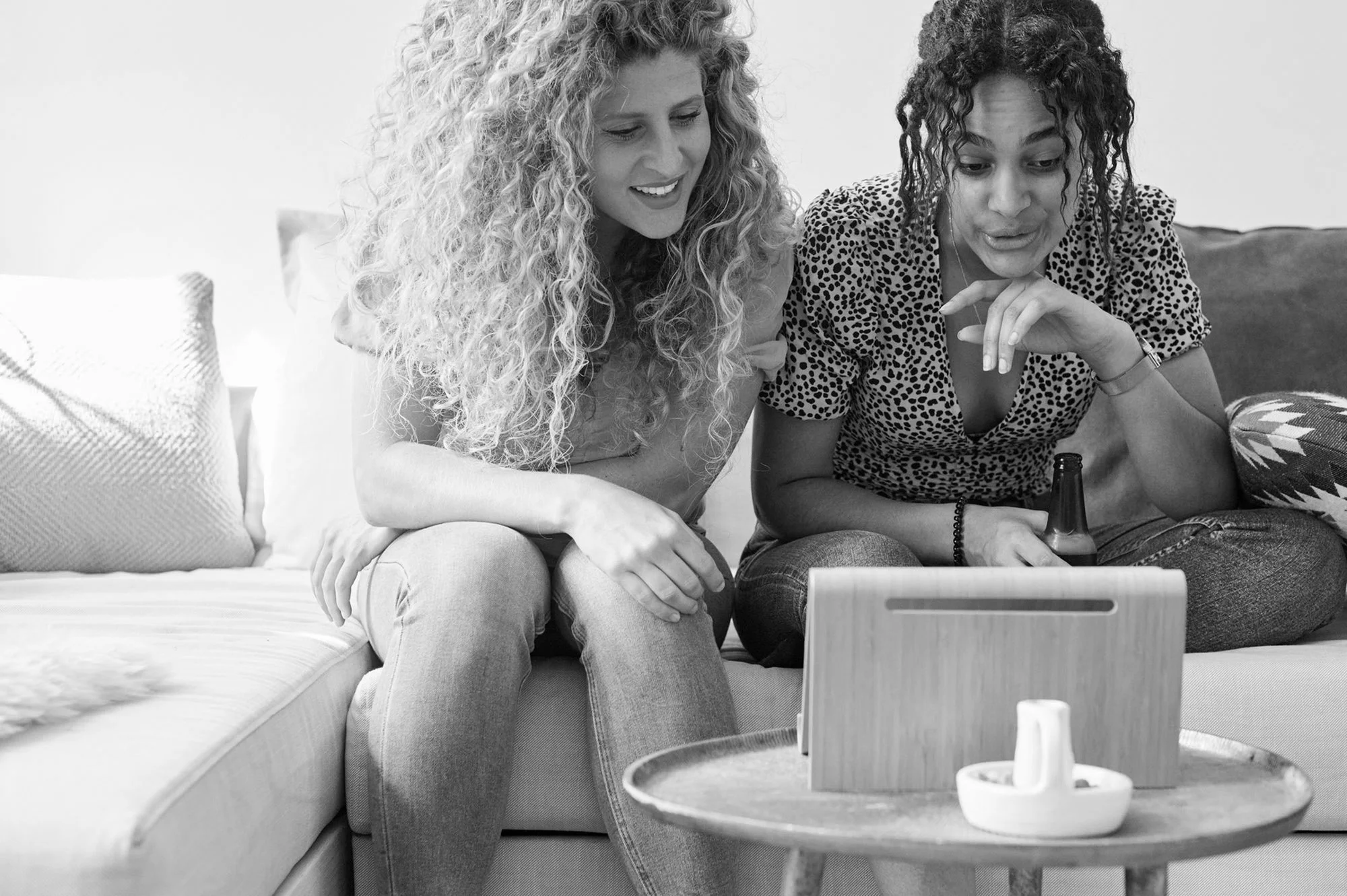 Two women sitting on a couch looking at a tablet, one holding a beer bottle, in a cozy living room.