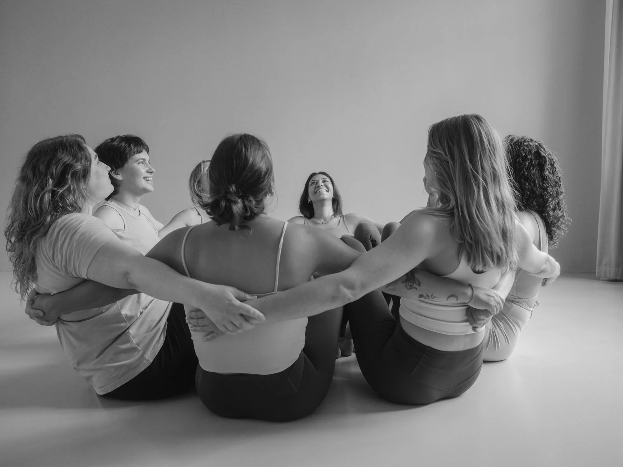 Group of women sitting on the floor in a circle holding hands and embracing, smiling and looking at each other in a joyful moment.