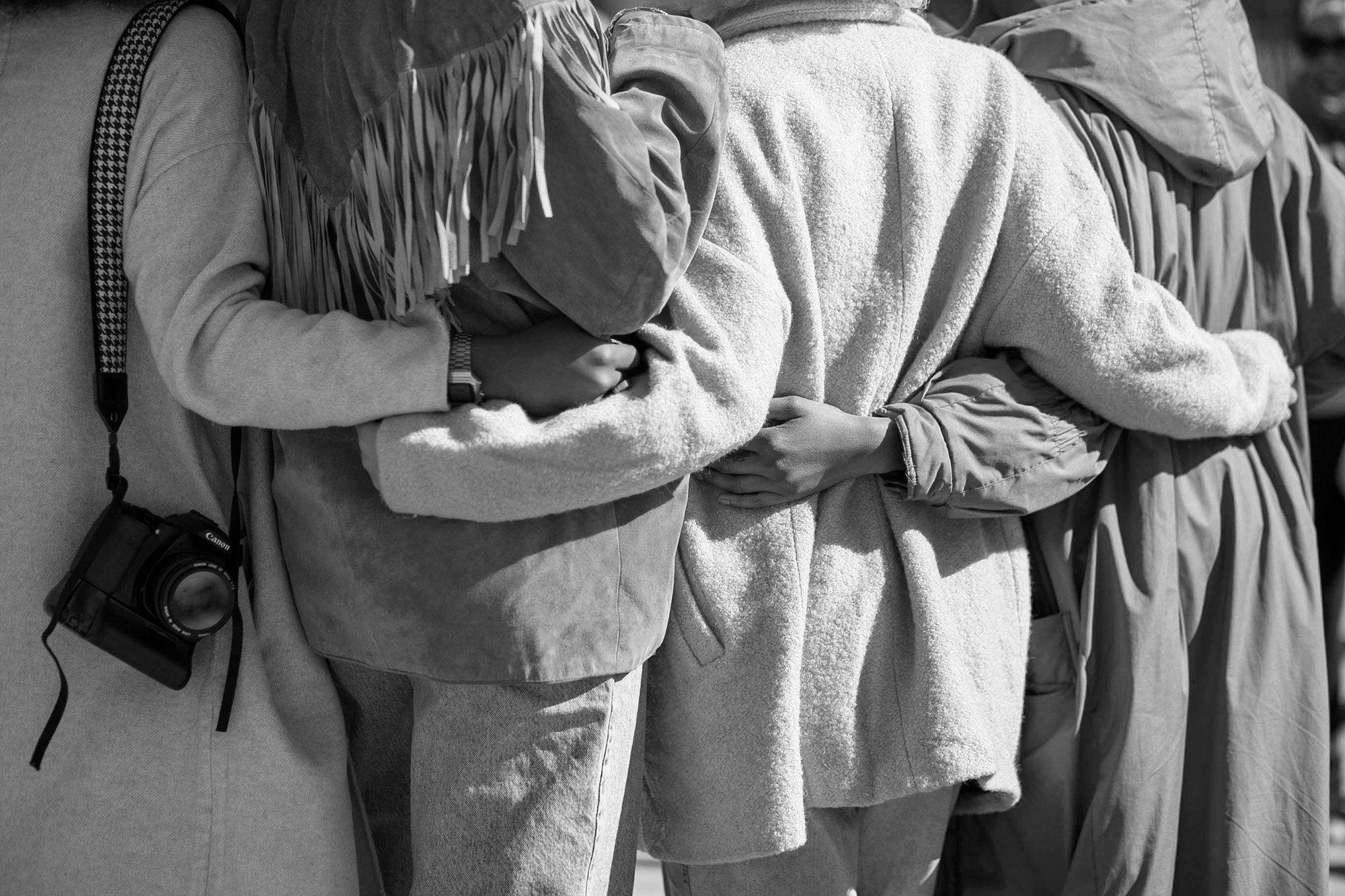 Three people standing in line, holding their stomachs, wearing jackets and coats. One person has a camera hanging from their neck.