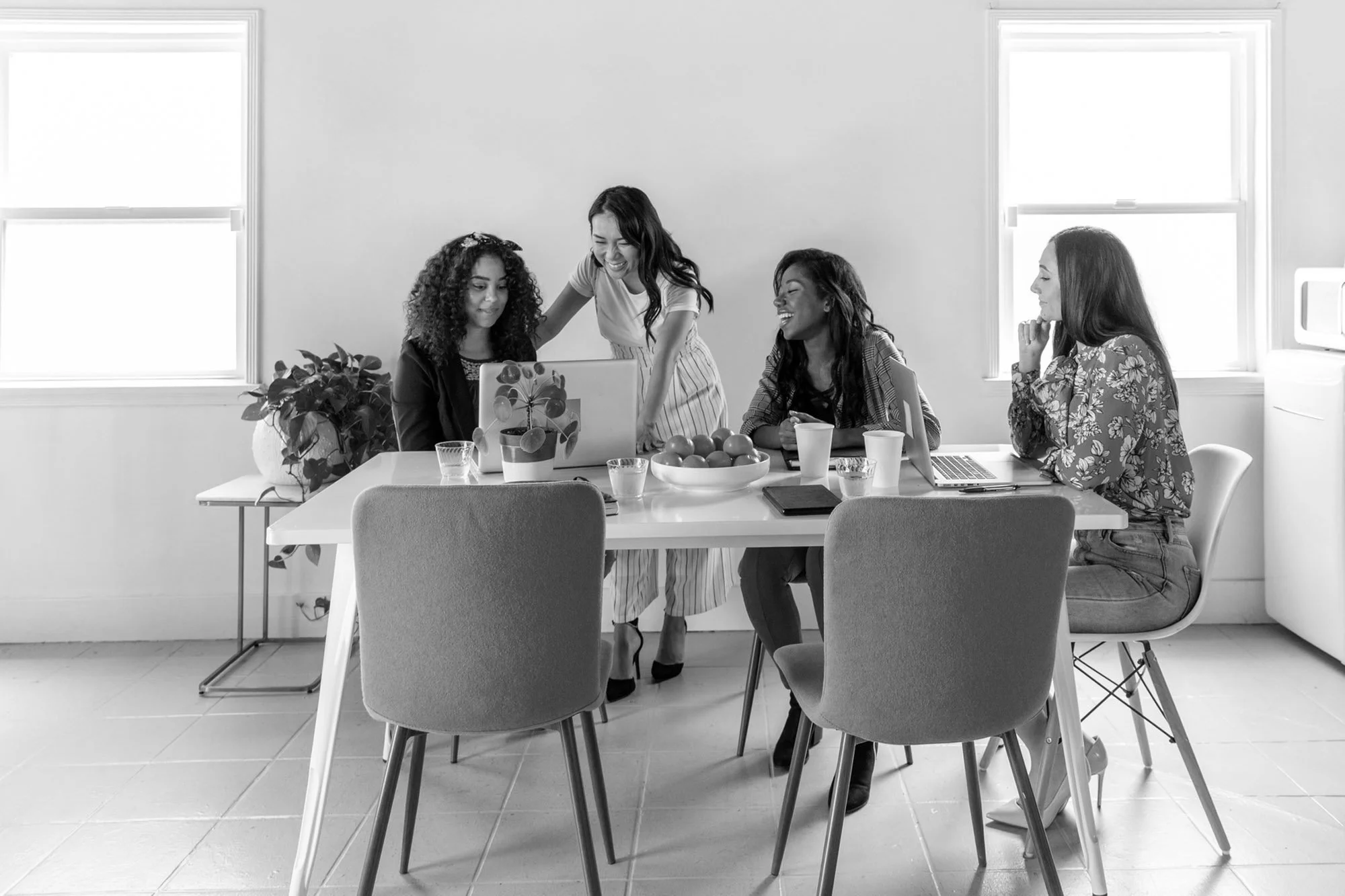 Four women gathered around a table in a bright room, looking at a laptop and smiling, with drinks and a bowl of fruit on the table.