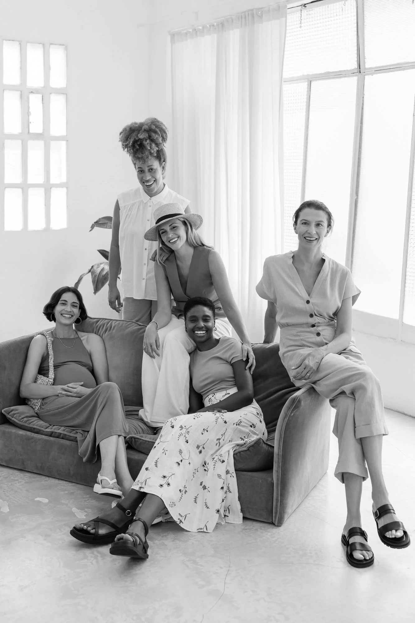 Five women smiling and sitting on and around a sofa in a well-lit room with large windows and curtains, black and white photograph.