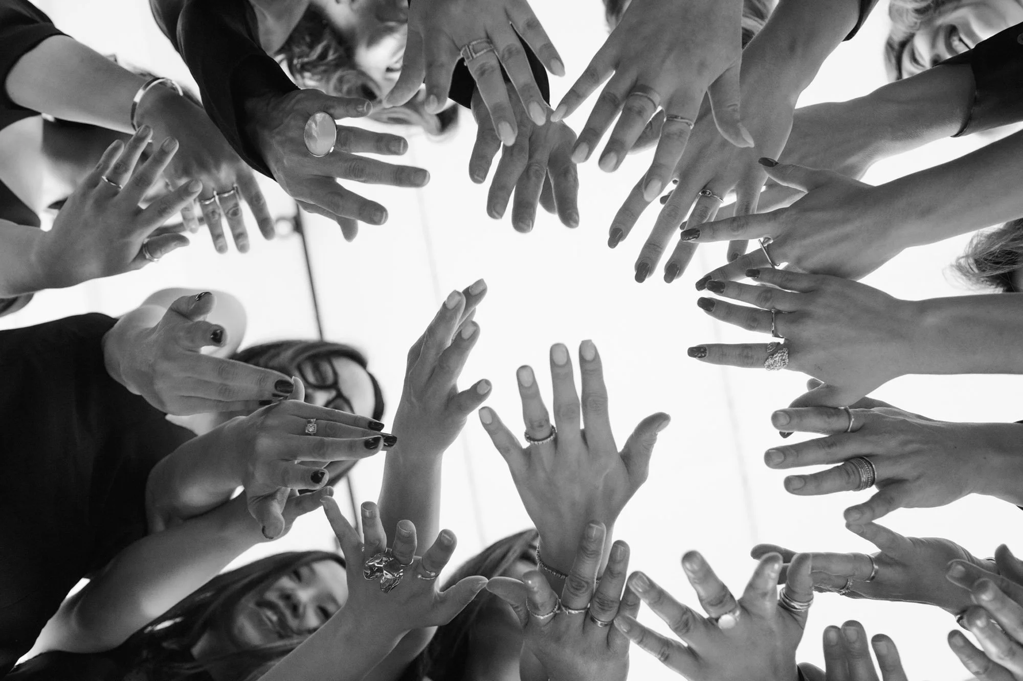 Black and white photograph of numerous hands reaching toward the center, showing diverse rings and nail polish, with a smiling woman visible in the background.