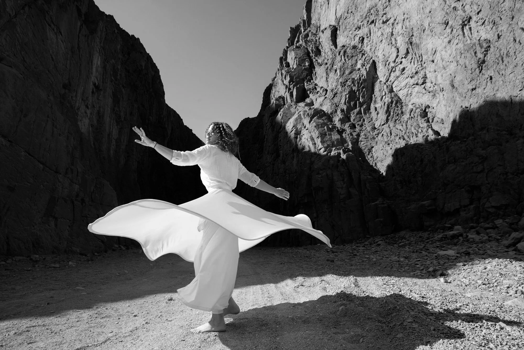 A woman in a white dress dancing in a canyon with rocky walls, her arms extended and her dress swirling as she twirls.