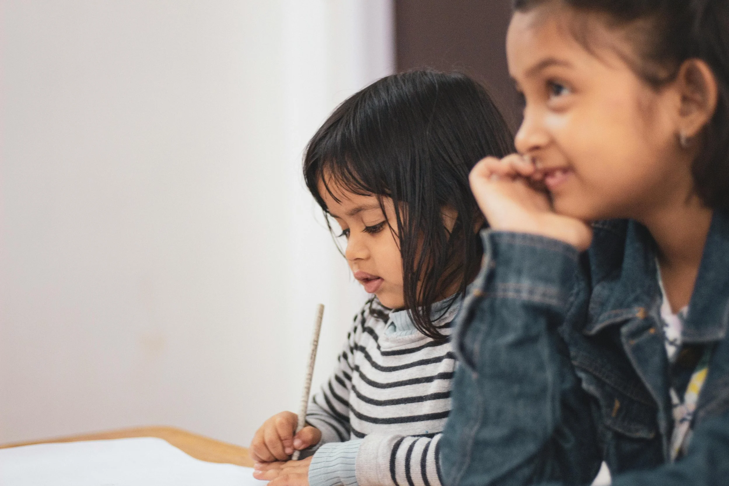 Two young girls sitting at a table, one writing with a pencil and the other resting her chin on her hand, smiling.