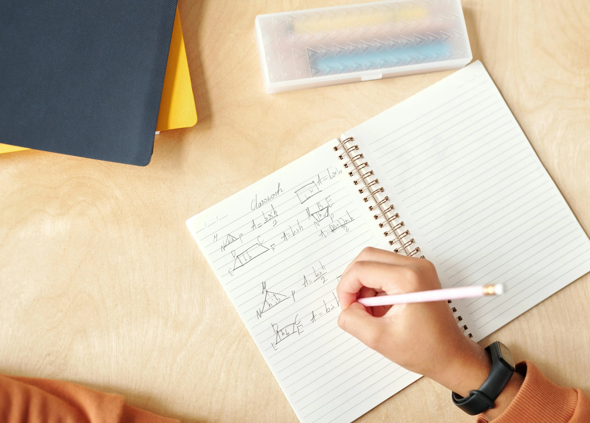 A person with a black watch on their wrist is writing physics notes with a pink pen in a spiral notebook on a wooden desk. There are two closed notebooks, one navy blue and one yellow, stacked on the upper left corner of the desk. A small transparent case with geometric patterns is placed above the notebook.