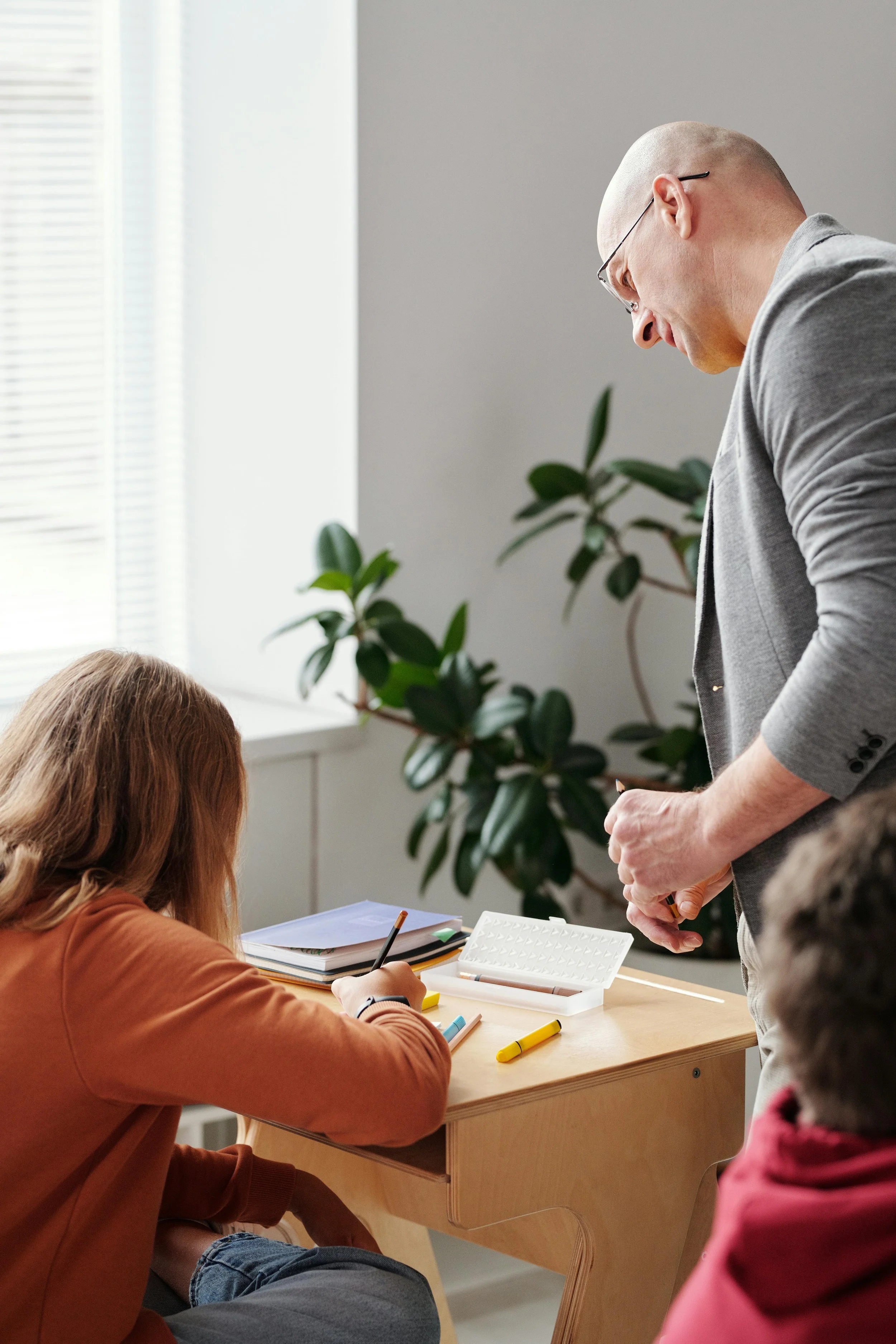 A teacher standing and talking to students sitting at a desk in a classroom, with notebooks and school supplies on the desk, and a large window with blinds and green plants in the background.