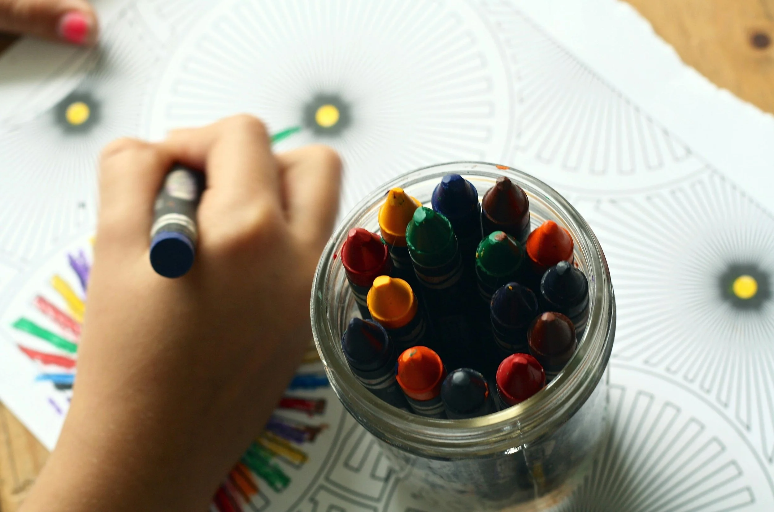 A person holding a blue marker and coloring on a color wheel chart, with a jar of assorted colored markers on the table.