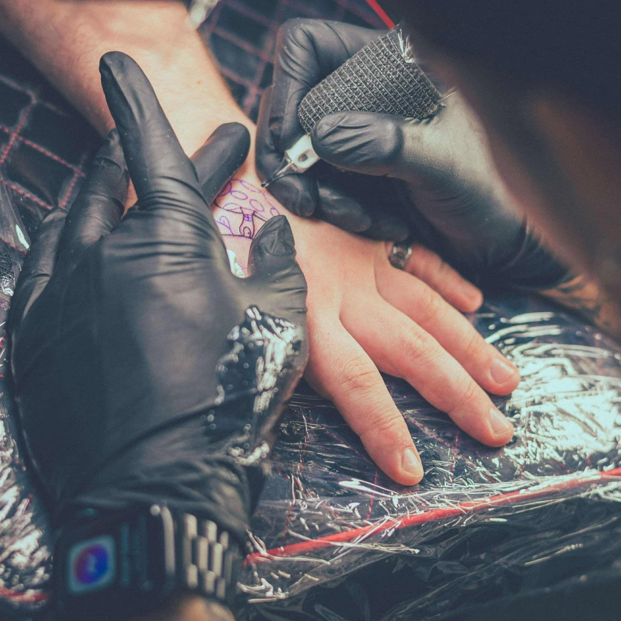 A person getting a tattoo on their hand by a tattoo artist wearing black gloves, with the tattoo machine inked on their hand and a protective plastic sheet covering the work area.