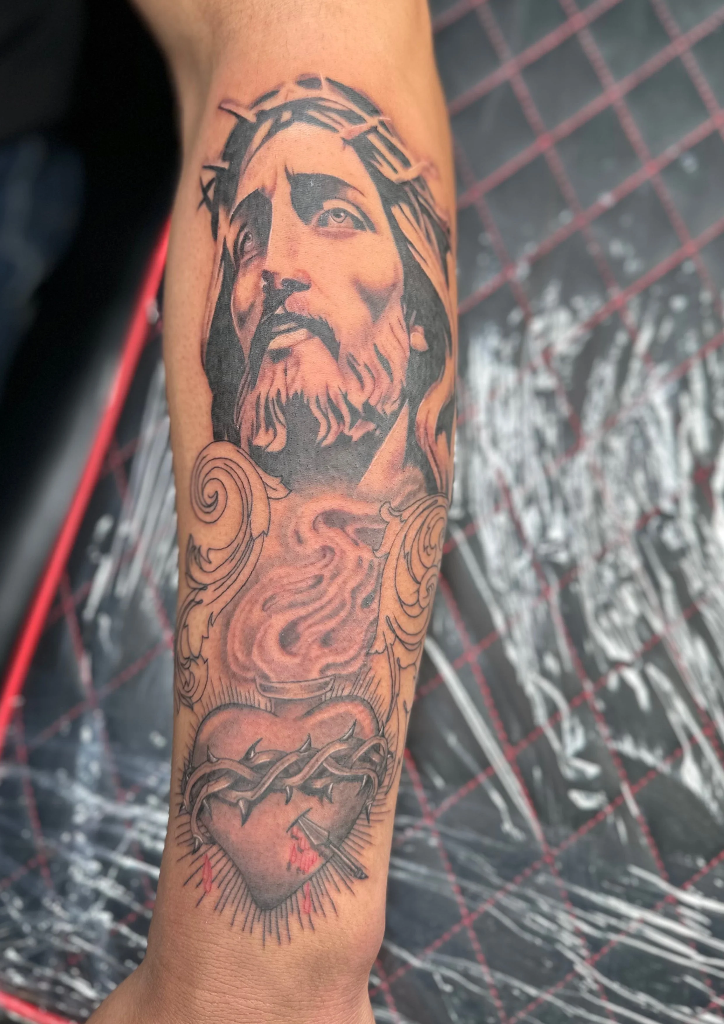 Tattoo of Jesus Christ wearing a crown of thorns, with flowing hair and beard, with a sacred heart surrounded by barbed wire and red drops below.