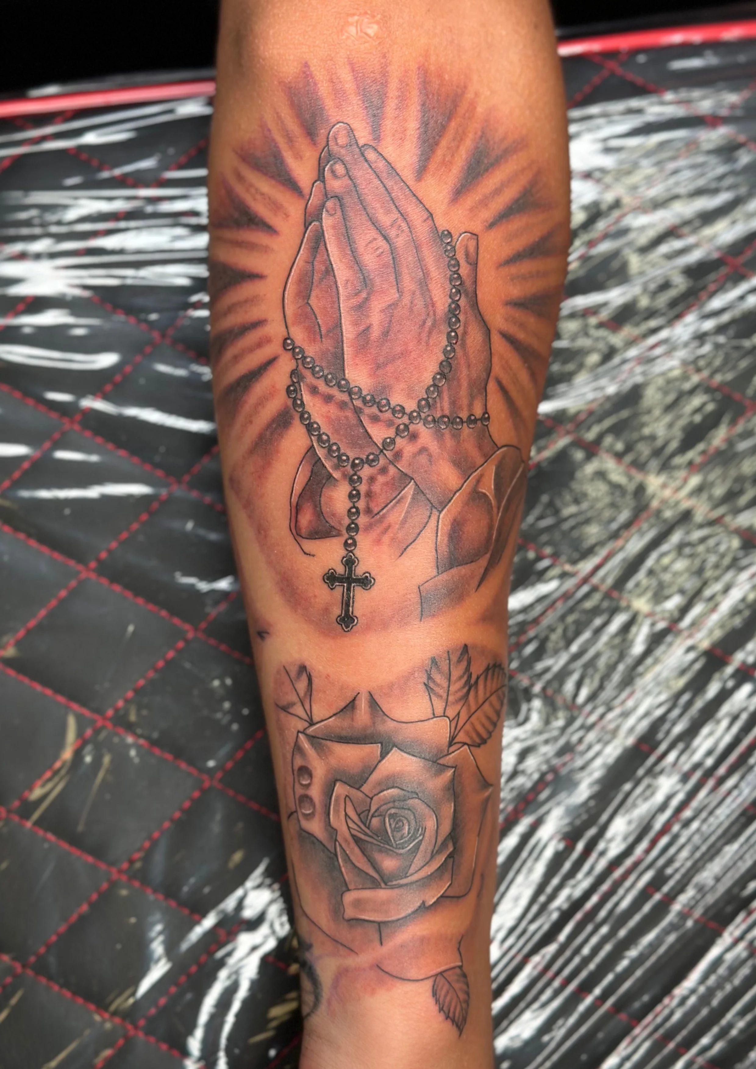 Tattoo of praying hands with a rosary, above a rose, on a person's forearm.