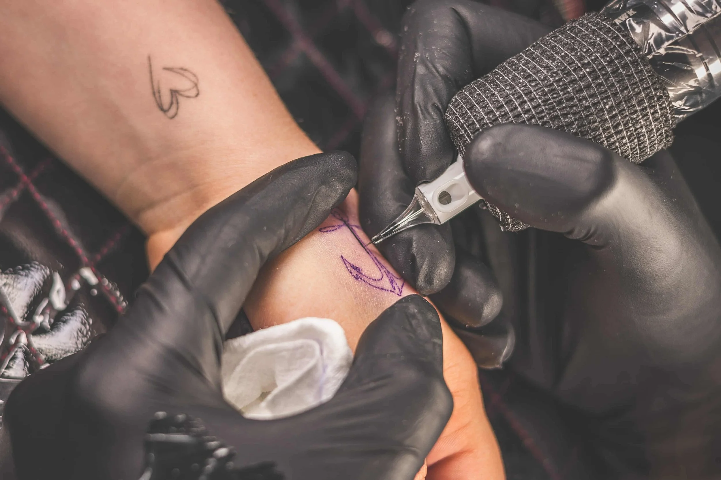 Tattoo artist wearing black gloves uses a needle to ink a purple tattoo design on a person's forearm.