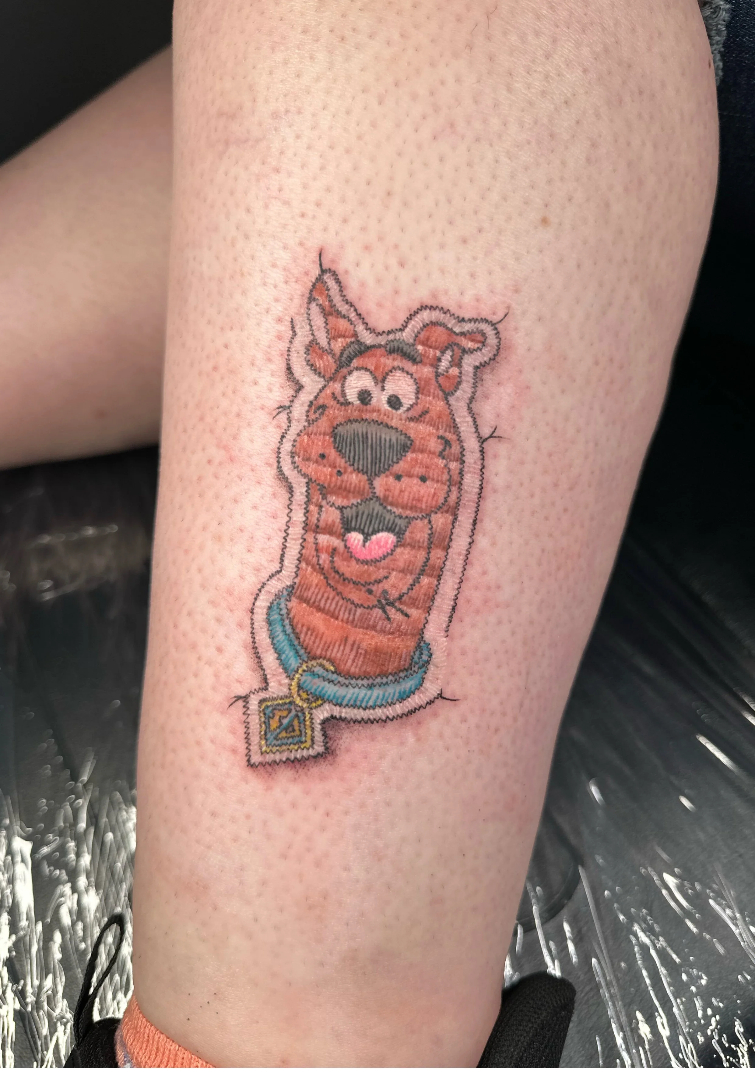 Colorful cartoon tiger tattoo on person's leg, featuring a collar with a pendant.
