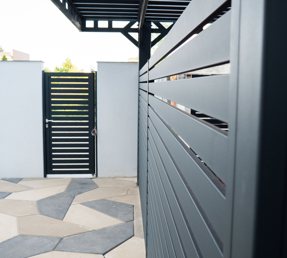 View of a black gate and fence, with a white wall and hexagonal stone pavement in the foreground.