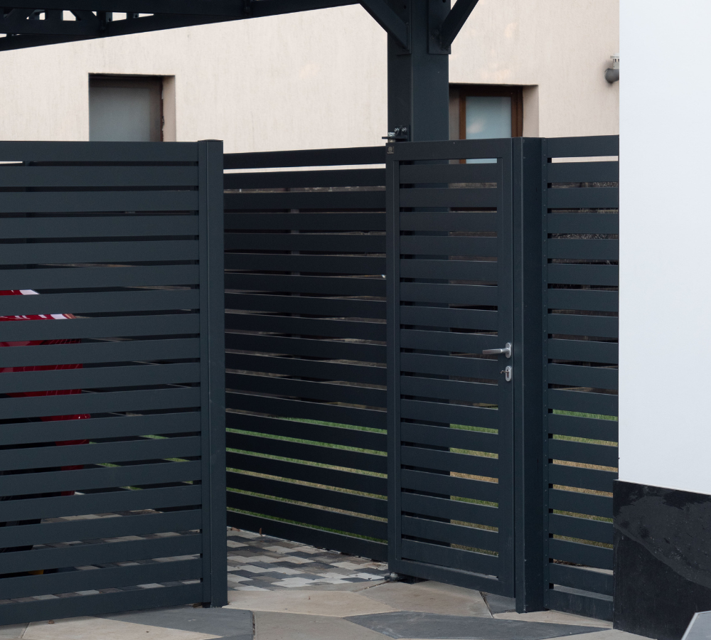 A black metal gate and fence in front of a light-colored building with two small windows. The gate is slightly open and is part of a modern, horizontal slat fencing design.