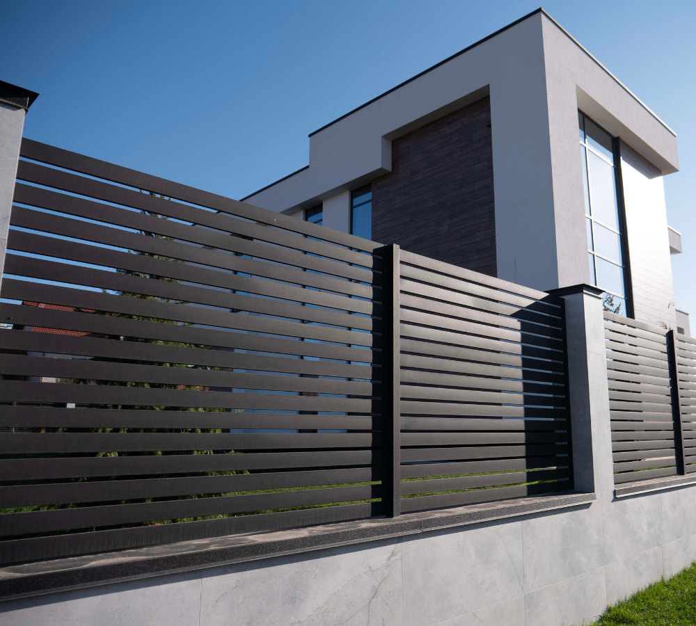 Modern house with a white exterior and large glass windows, surrounded by a black horizontal slat fence, under a clear blue sky.