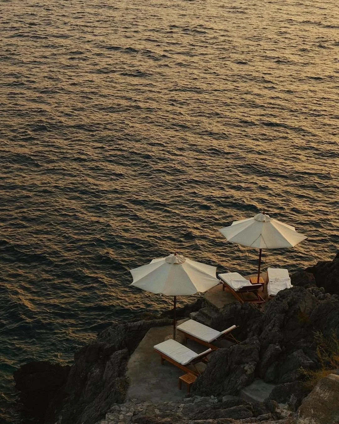 Aerial view of two white sun loungers and umbrellas on a private rocky platform overlooking the dark Mediterranean sea at sunset. A secluded spot for slow living in Italy.
