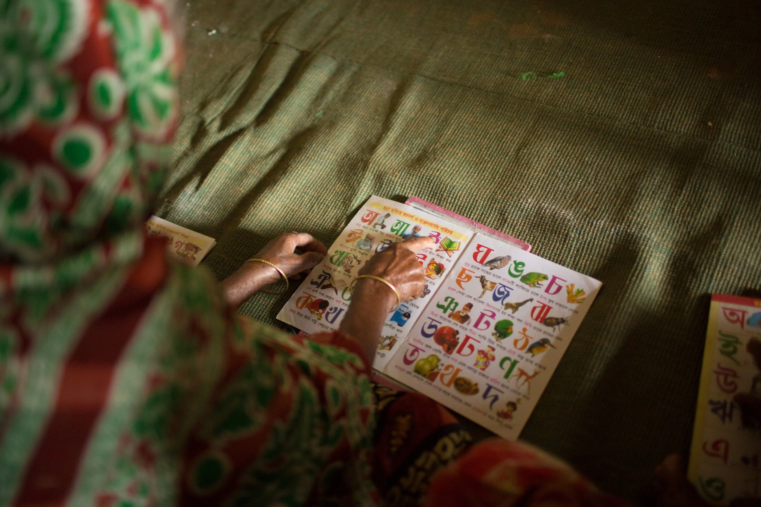A person sitting on a mat, wearing a colorful patterned saree, looking at children’s alphabet flashcards with illustrations in Bengali.