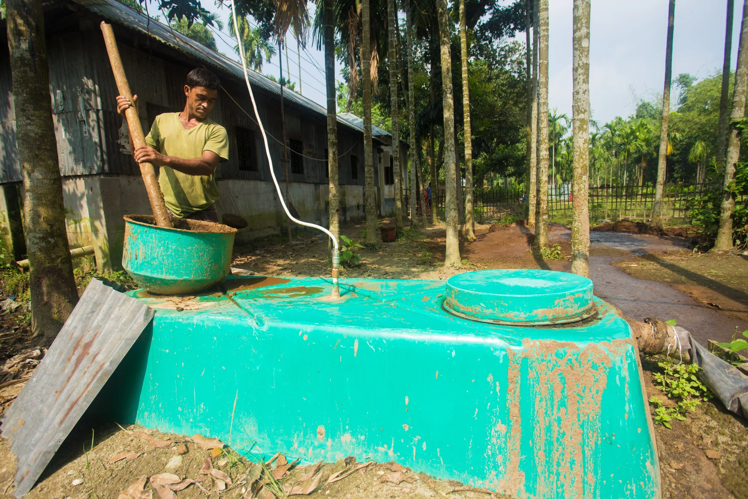 Man operating a biogas tank outdoors among trees and a rustic building. Generating clean energy from compost and agricultural byproducts to reduce fuel use and improve air quality.