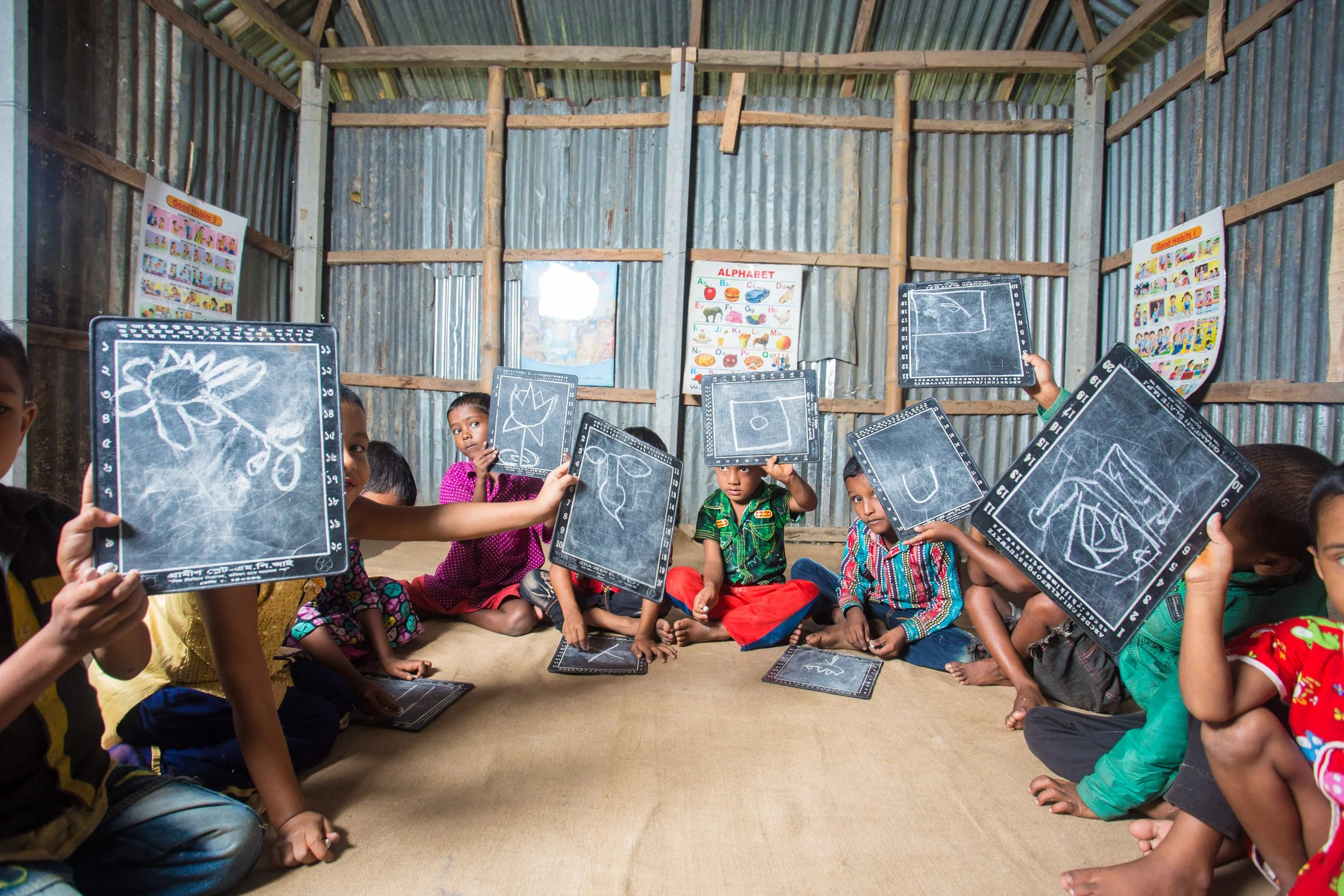 Children sitting on the floor inside a rustic classroom, holding up chalkboard tablets with drawings, in front of colorful educational posters on the walls.