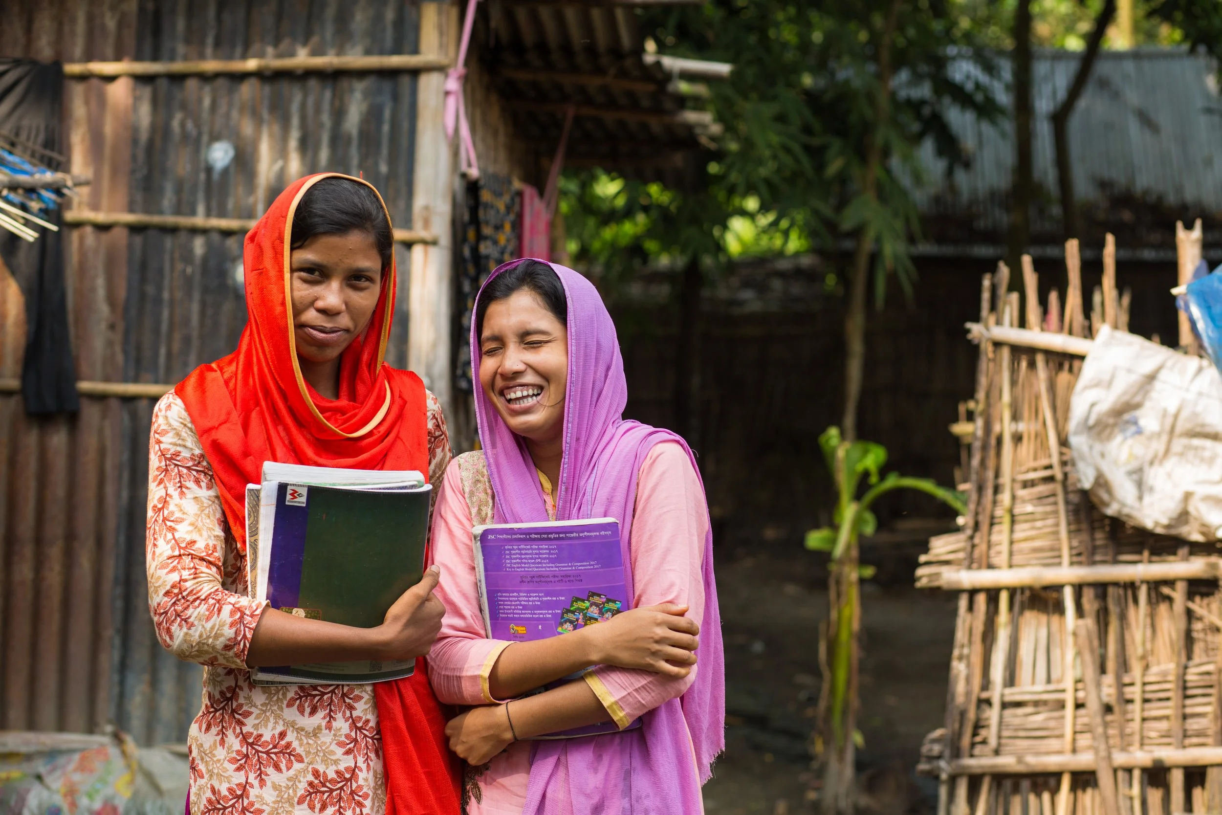 Two women in colorful traditional clothing standing outside, holding notebooks and smiling, benefactors of women's education at Kazi and Kazi organic tea garden.
