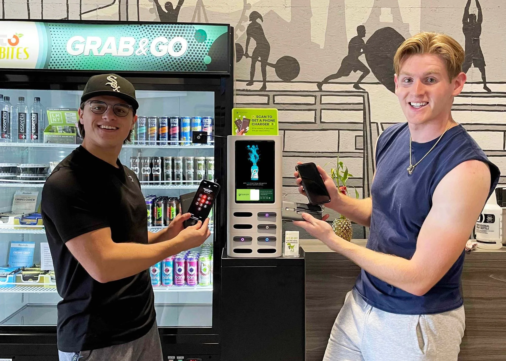 Two young men using their phones to pay at a vending machine inside a store, behind them a refrigerator stocked with drinks, and a wall with artistic sports figures and equipment.