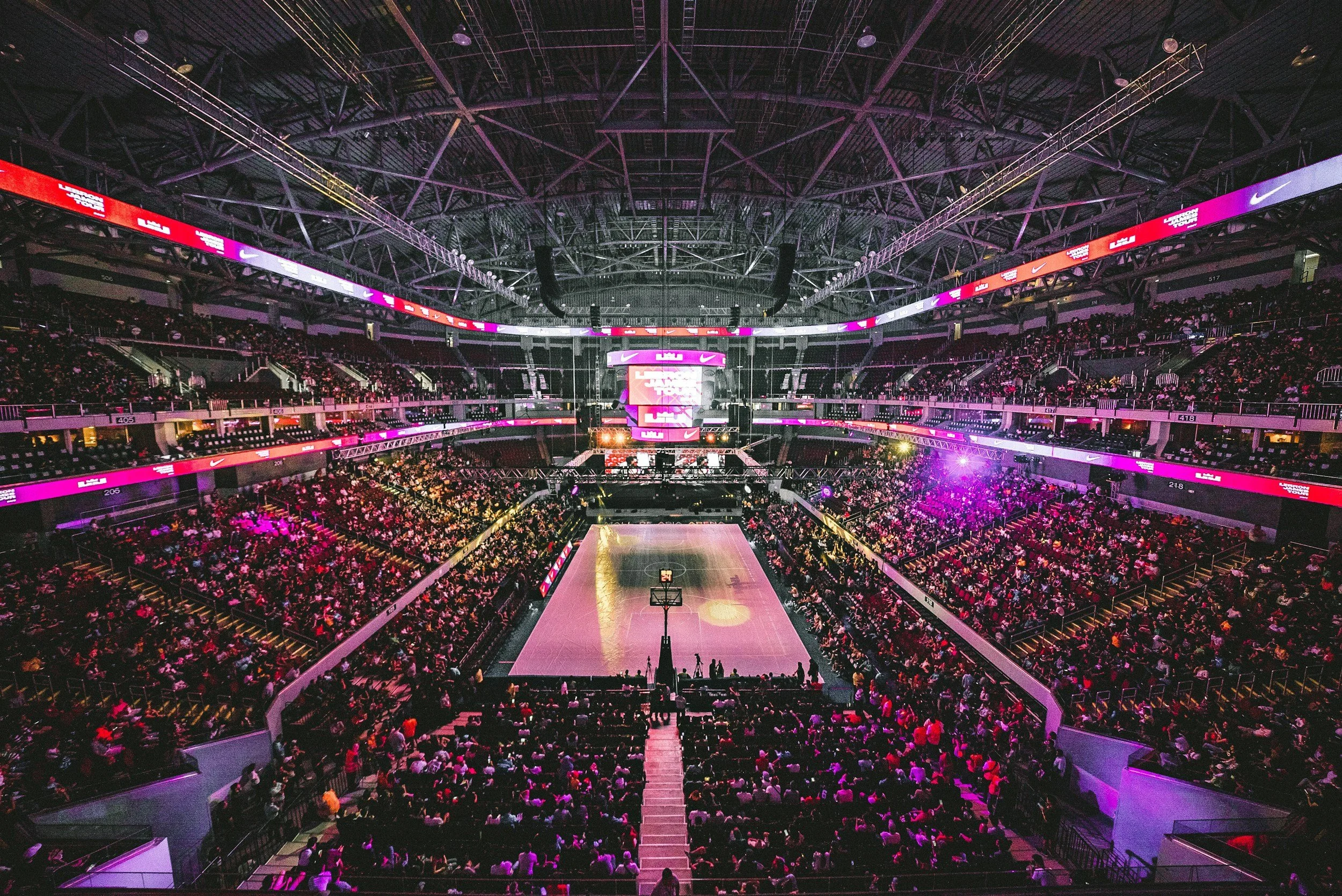 Interior view of a basketball arena filled with spectators, with the court lit up and large screens hanging above.