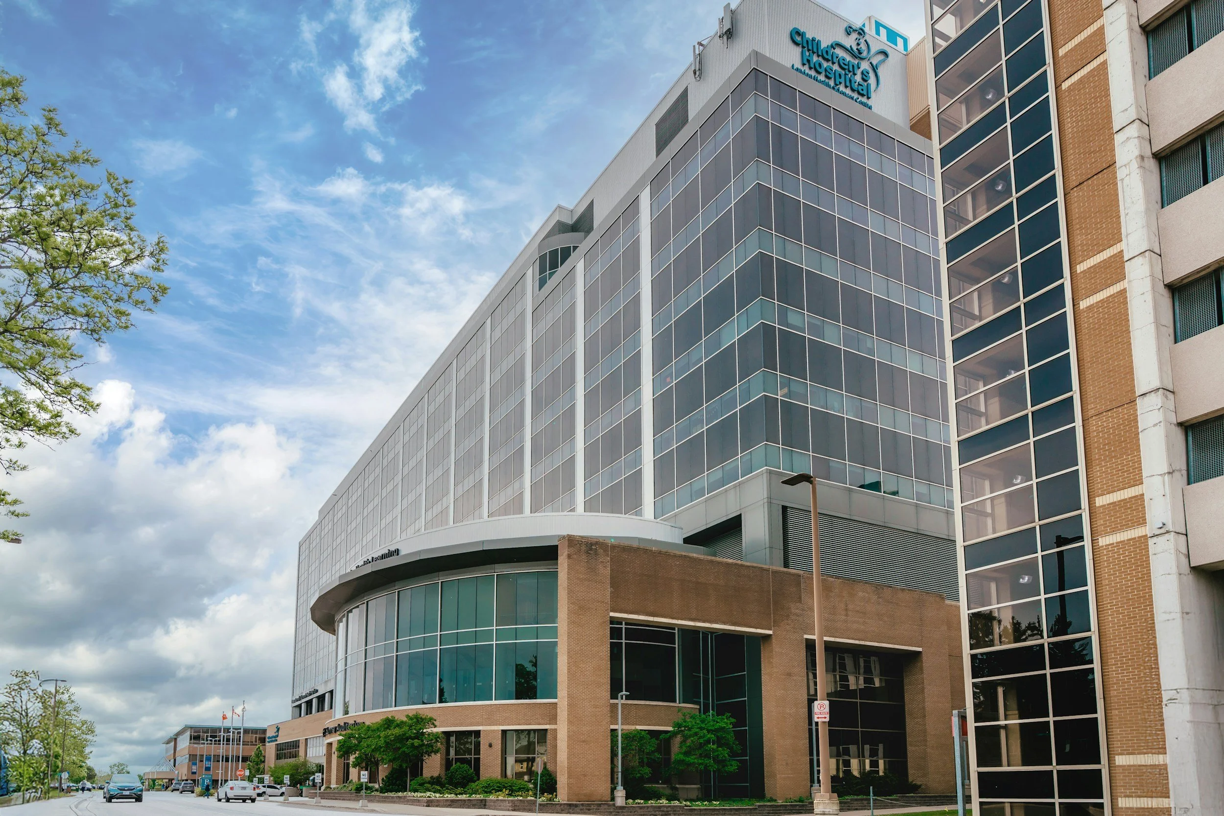Exterior view of a modern hospital building with glass windows, situated on a city street with cars and green trees.