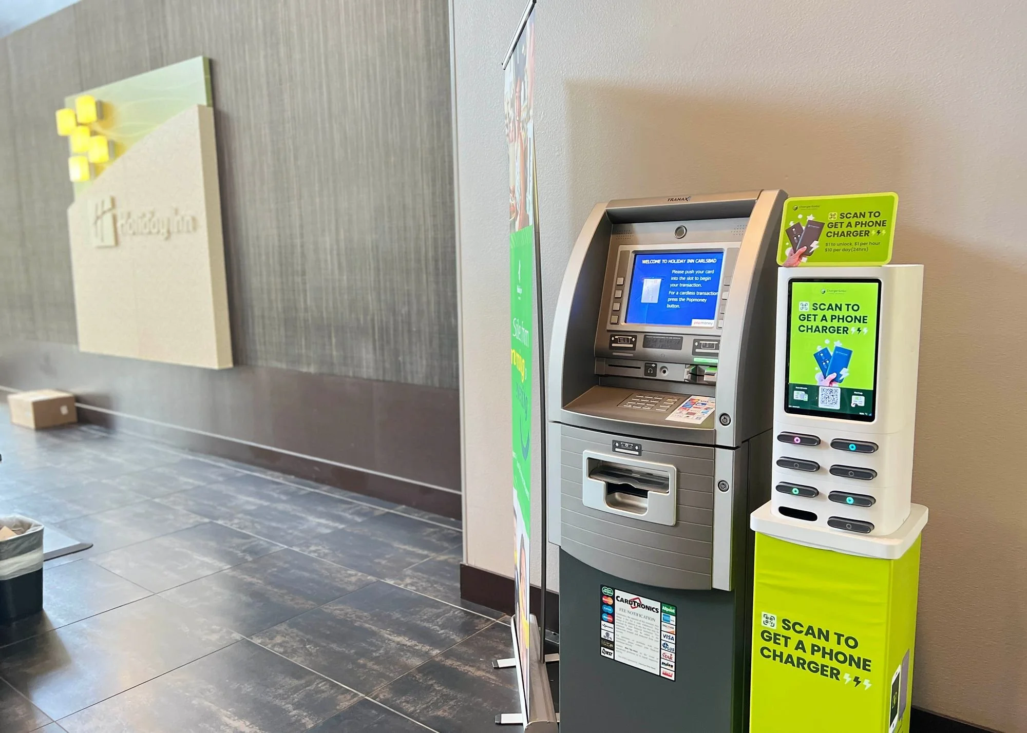 An ATM machine and a phone charger kiosk at a hotel lobby. The ATM has a blue screen with instructions, and the kiosk has a bright green sign offering to scan a QR code to get a phone charger.