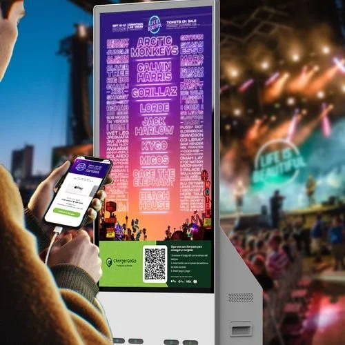 A person purchasing a ticket from a digital kiosk at an outdoor music festival. The screen displays a lineup including Arctic Monkeys, Calvin Harris, Gorillaz, and others, with a sunset background and festival crowd in the distance.