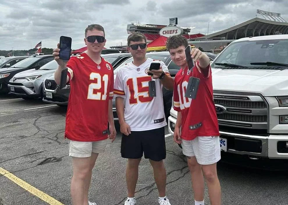 Three young men standing in a parking lot, holding up smartphones, with a stadium in the background. They are wearing football jerseys and shorts.