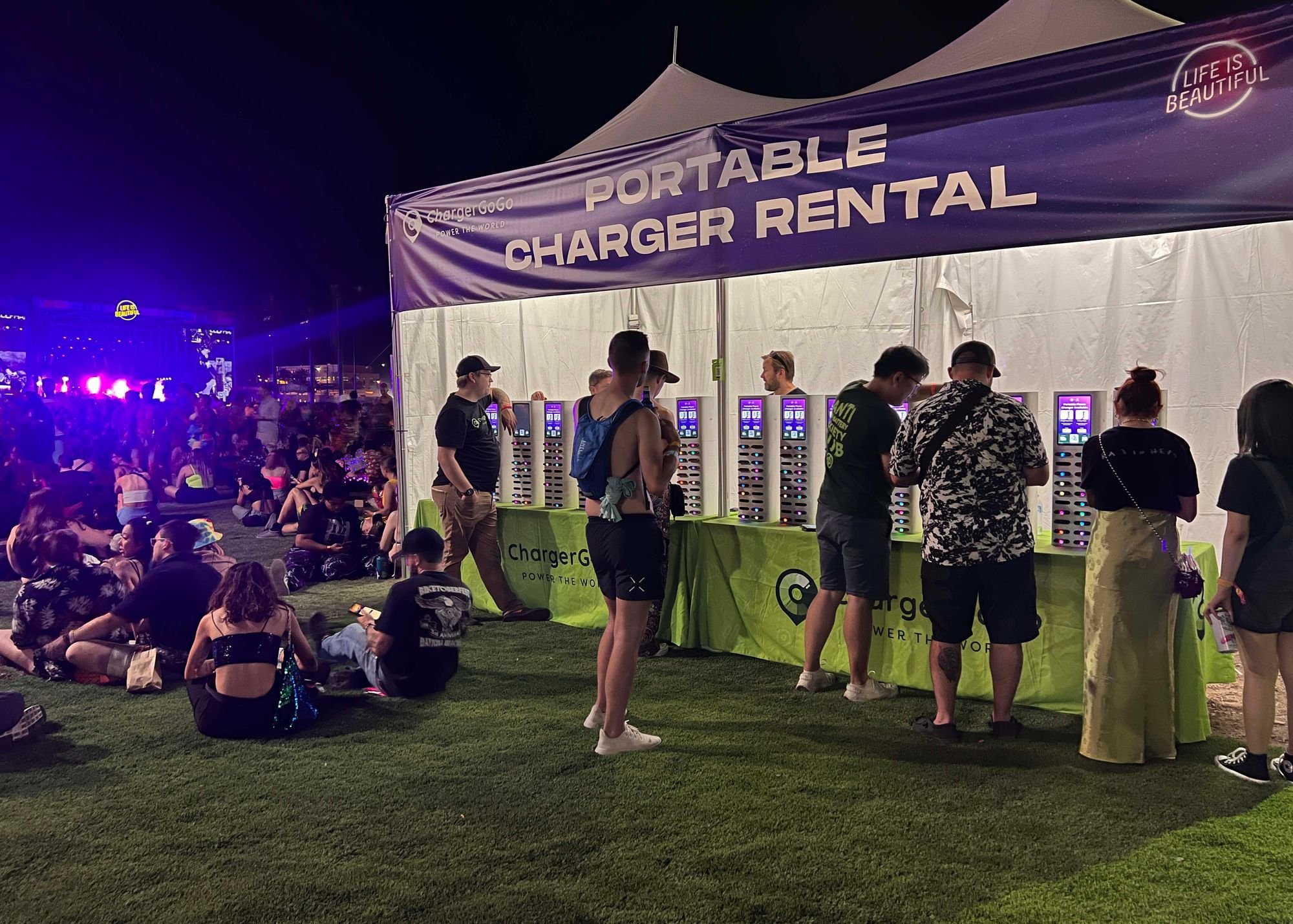 Crowd sitting on grass at an outdoor event at night, with a large tent labeled 'Portable Charger Rental', and people using charging stations inside.