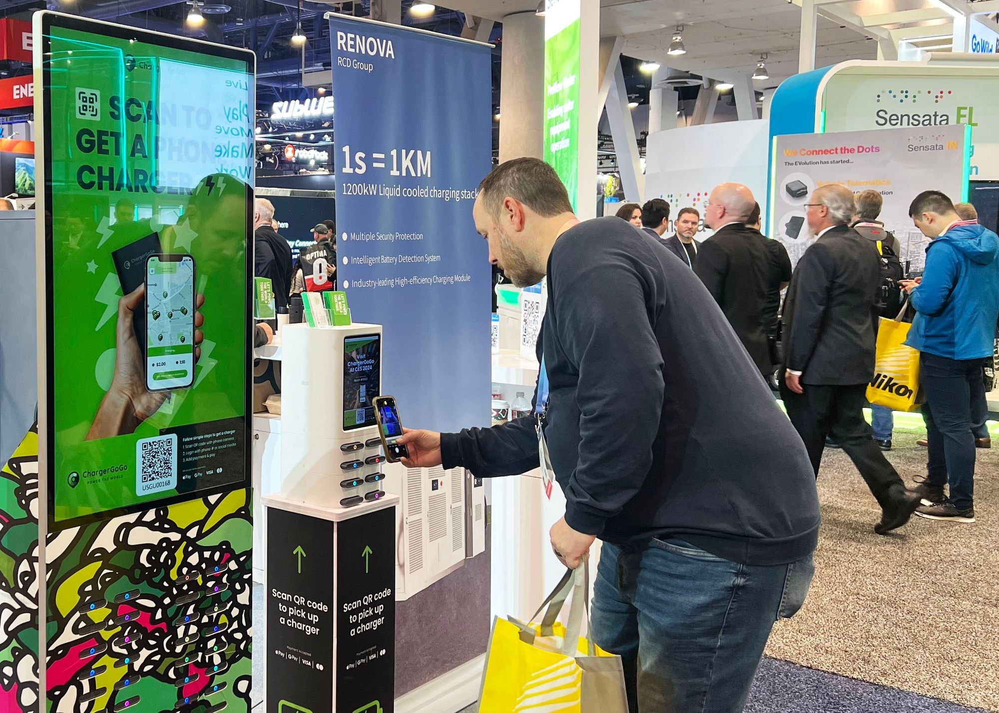 A man at a trade show scans a QR code on a white charging station with his smartphone. In the background, multiple people walk past various booths with signs detailing technology and energy solutions, including one that advertises a 1200KW liquid cooled charging station.
