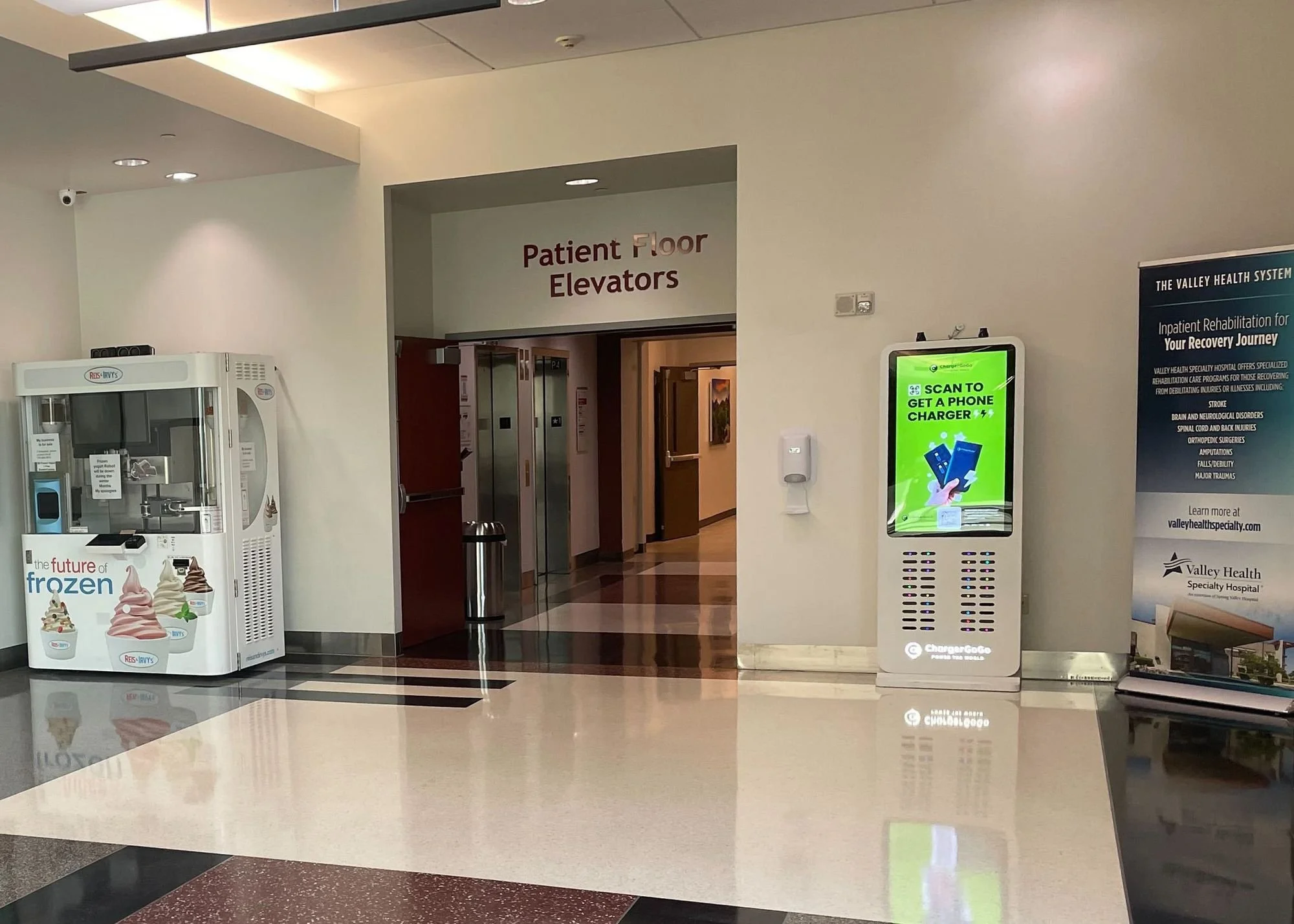 Hospital corridor with a sign reading 'Patient Floor Elevators,' a vending machine for frozen treats on the left, a digital signage for phone charging on the right, and a blue Valley Health System promotional poster on the far right.