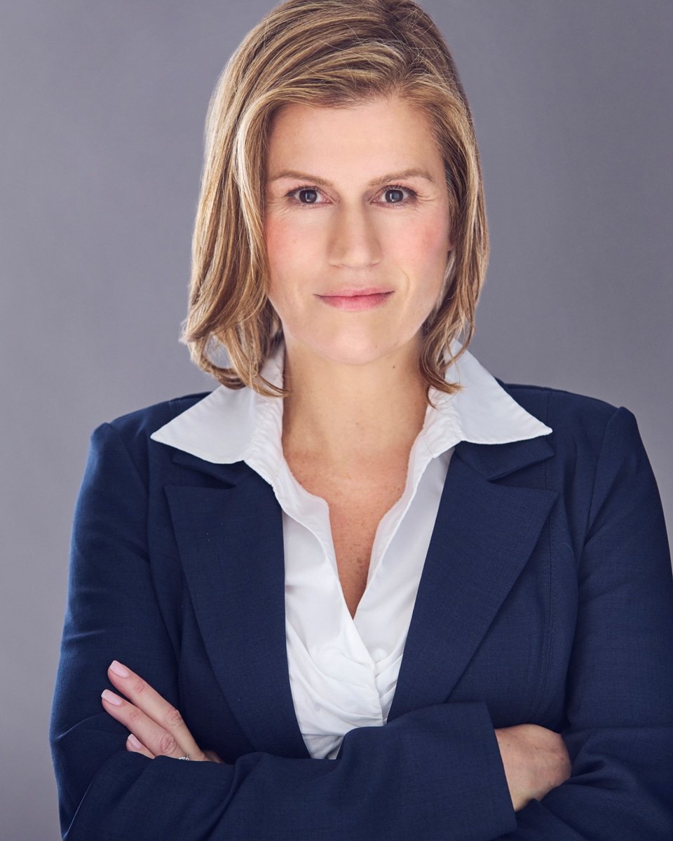 Headshot of comedian and actress Kerri Louise with shoulder-length blonde hair wearing a navy blazer over a white collared shirt, standing with arms crossed against a gray background.