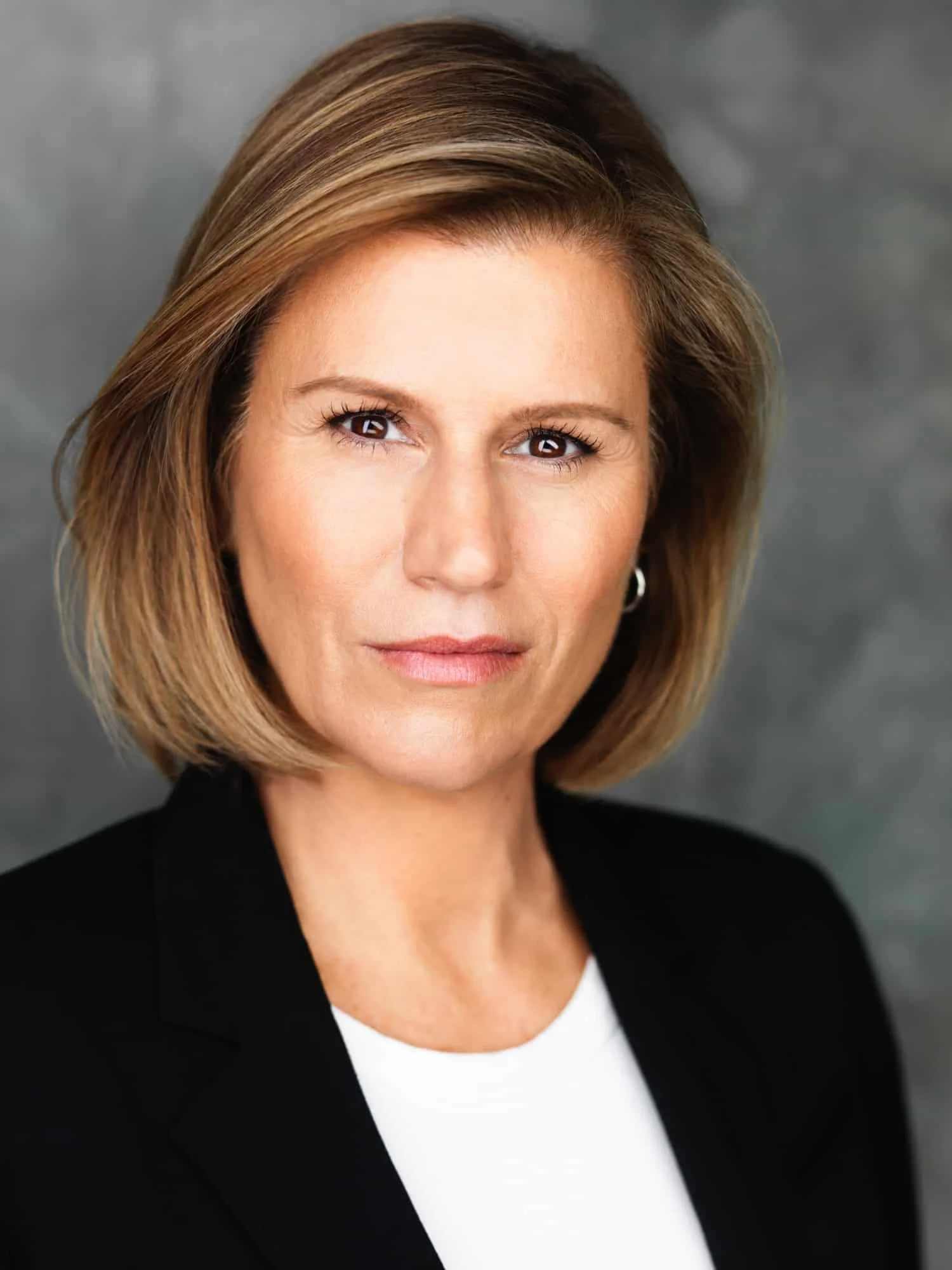 Headshot of comedian and actress Kerri Louise with blonde hair wearing a black blazer and white top against a gray background.