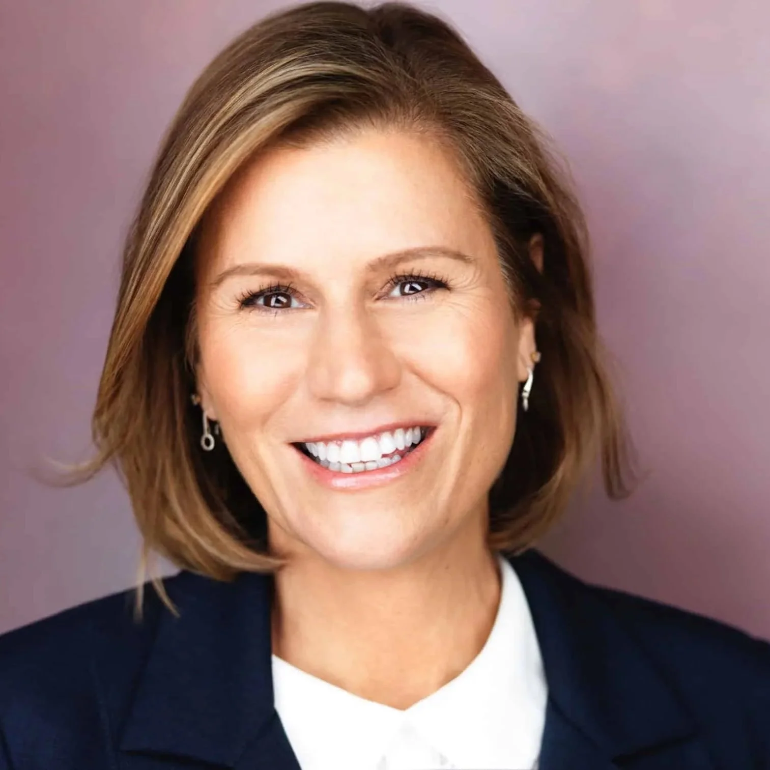 Close-up portrait of comedian and actress Kerri Louise, wearing earrings and a dark blazer against a purple background.