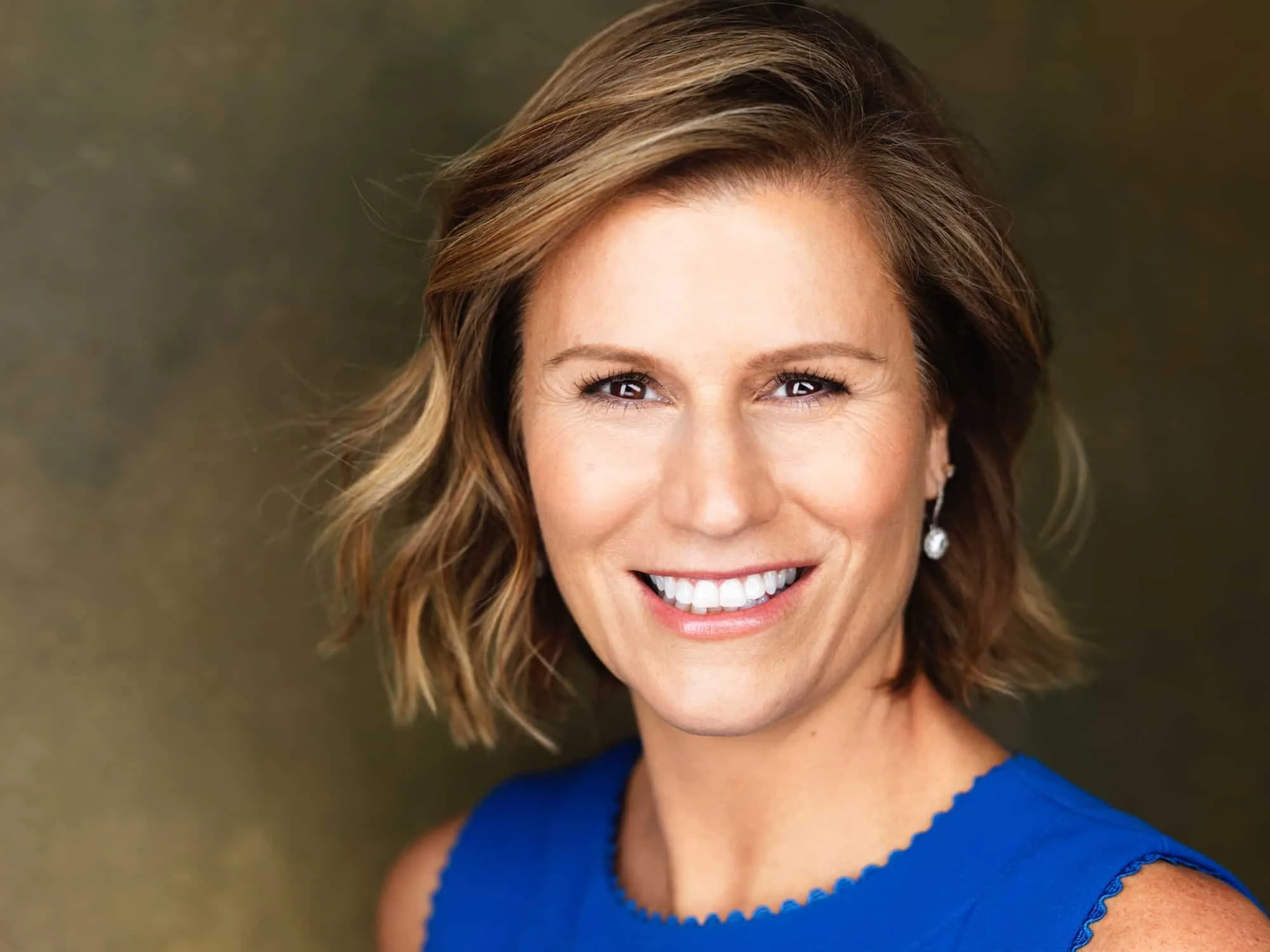 Headshot of comedian and actress Kerri Louise shoulder-length wavy light brown hair, blue eyes, and diamond earrings, wearing a sleeveless royal blue top, against a blurred outdoor background.