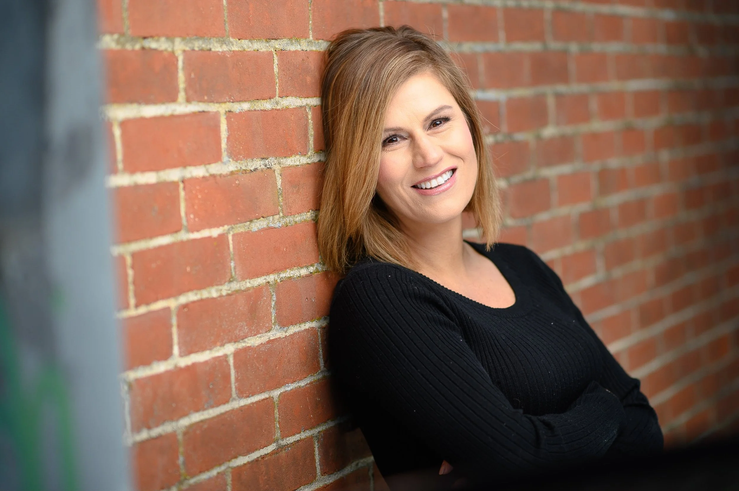 Comedian and actress Kerri Louise with shoulder-length light brown hair, smiling, leaning against a red brick wall, wearing a black long-sleeve top.