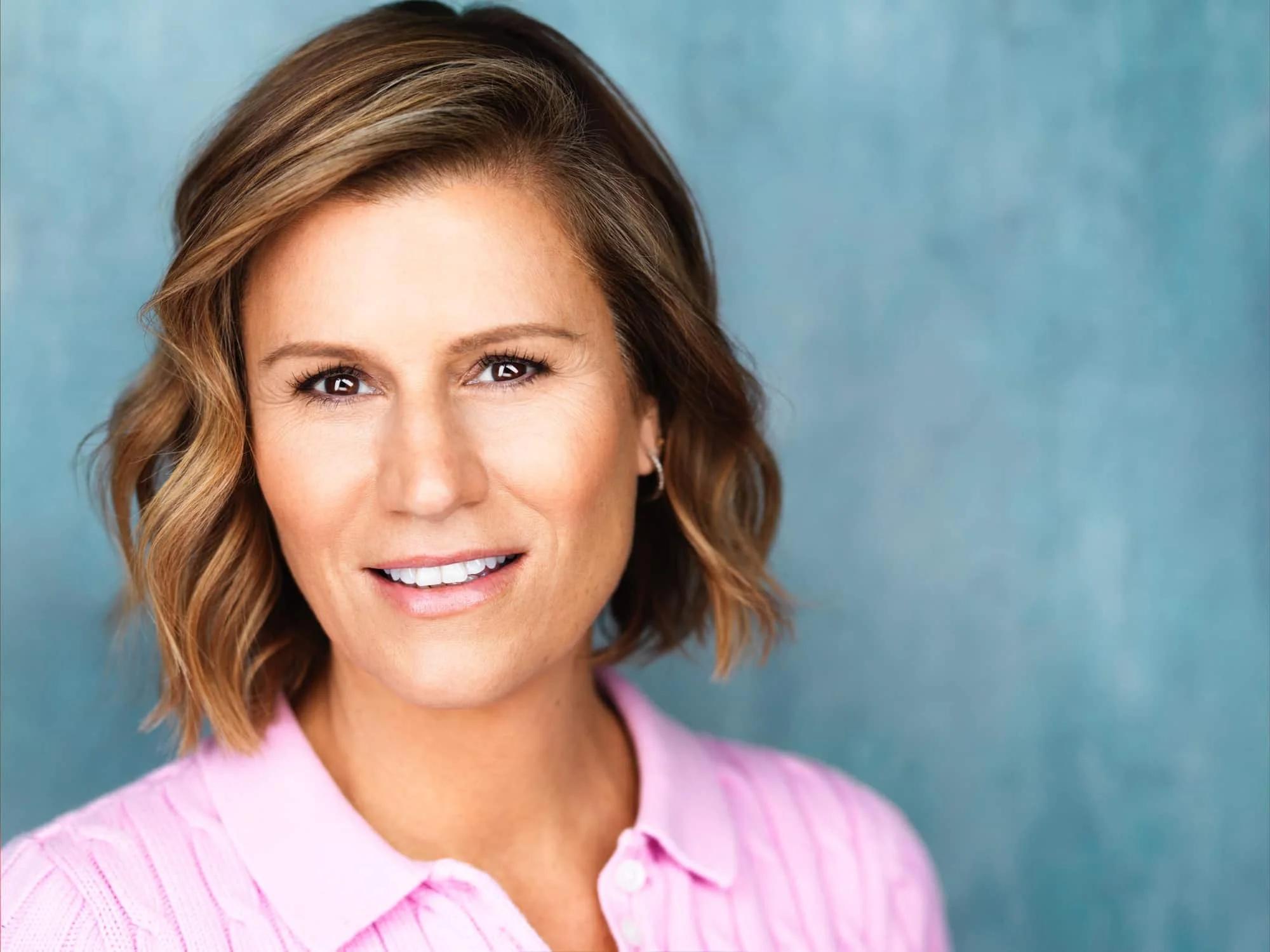Close-up portrait of comedian and actress Kerri Louise with short, wavy brown hair, smiling, wearing a pink collared shirt, against a blue background.