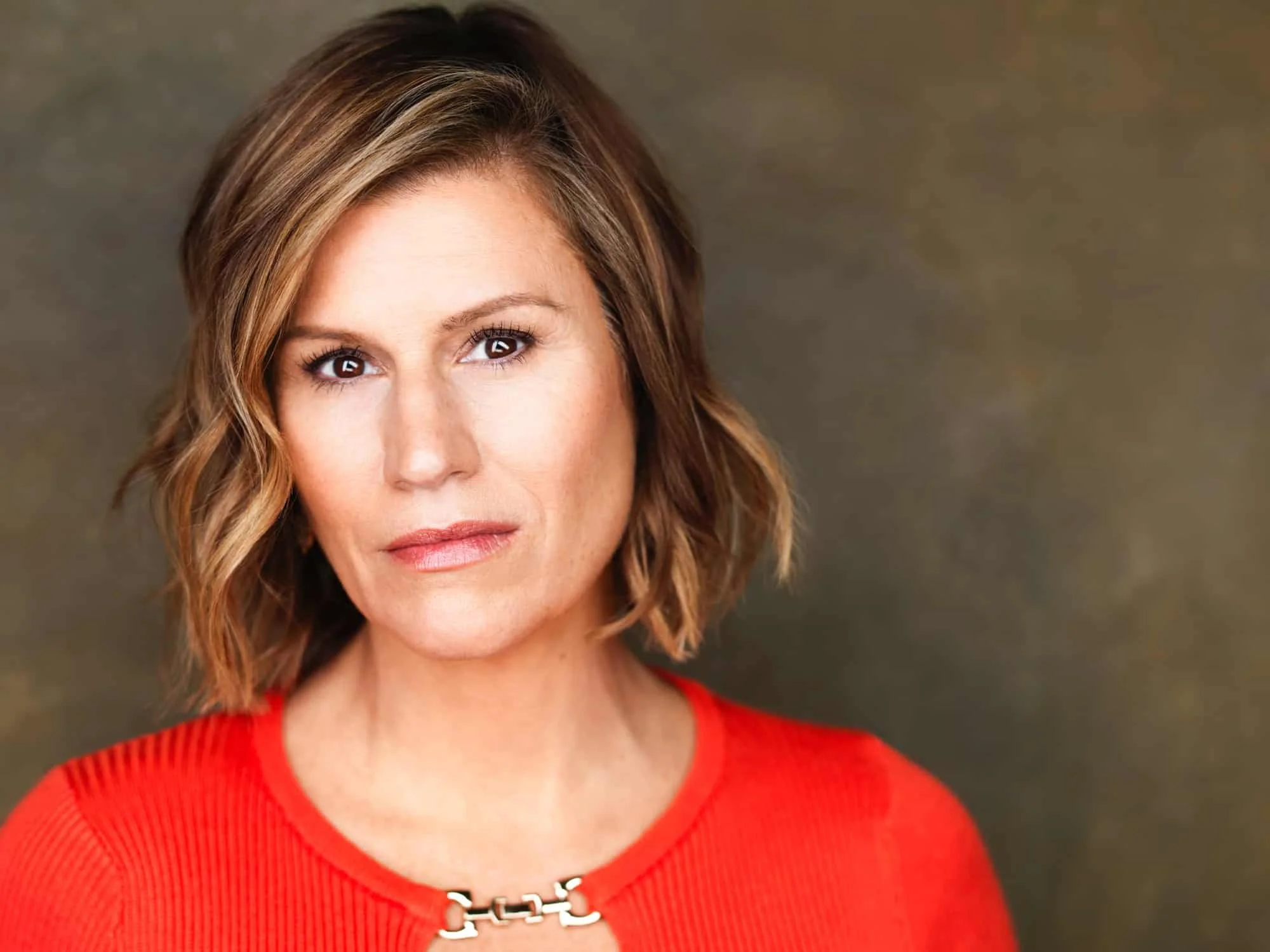 Headshot of comedian and actress Kerri Louise with short, wavy brown hair, wearing a red top and a silver chain necklace, looking directly at the camera.