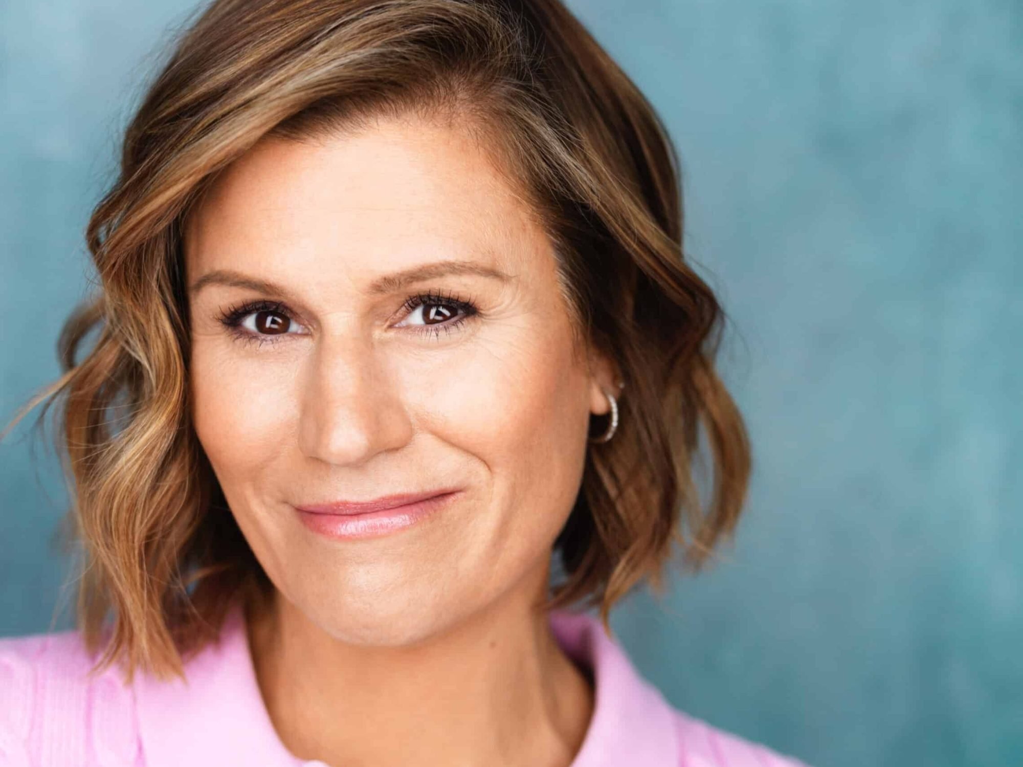 Headshot of comedian and actress Kerri Louise with short wavy brown hair, wearing hoop earrings and a pink top, against a blue background.