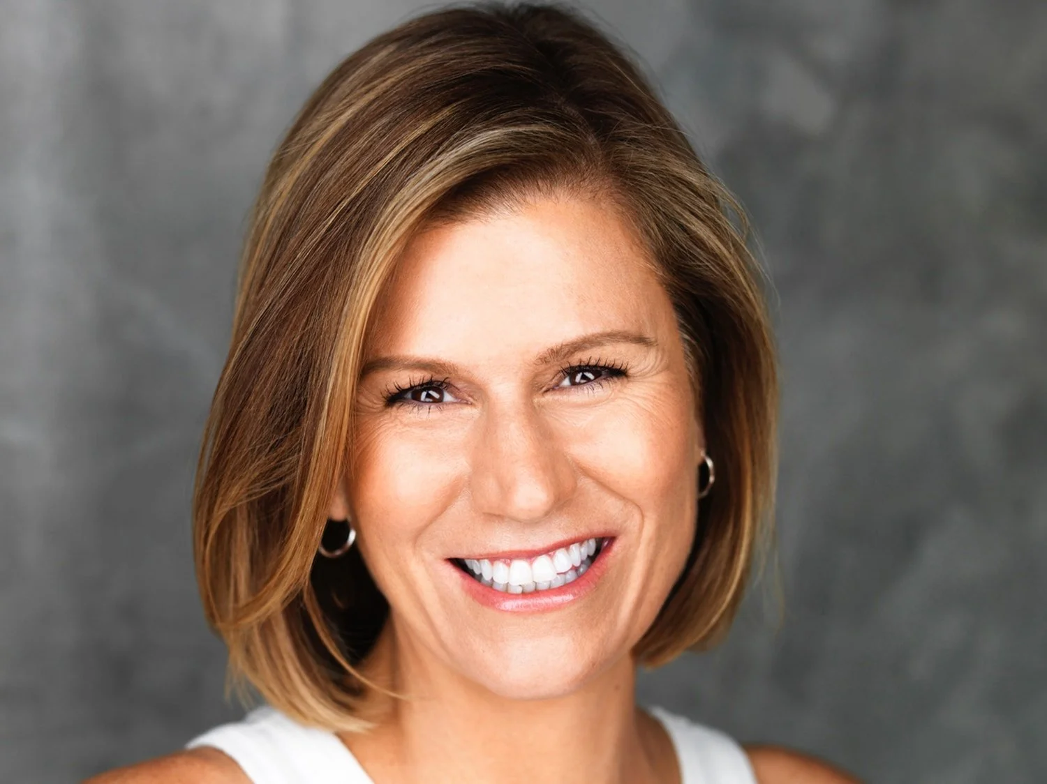 Comedian and actress Kerri Louise with shoulder-length light brown hair, smiling with white teeth, wearing hoop earrings and a white top, against a gray textured background.