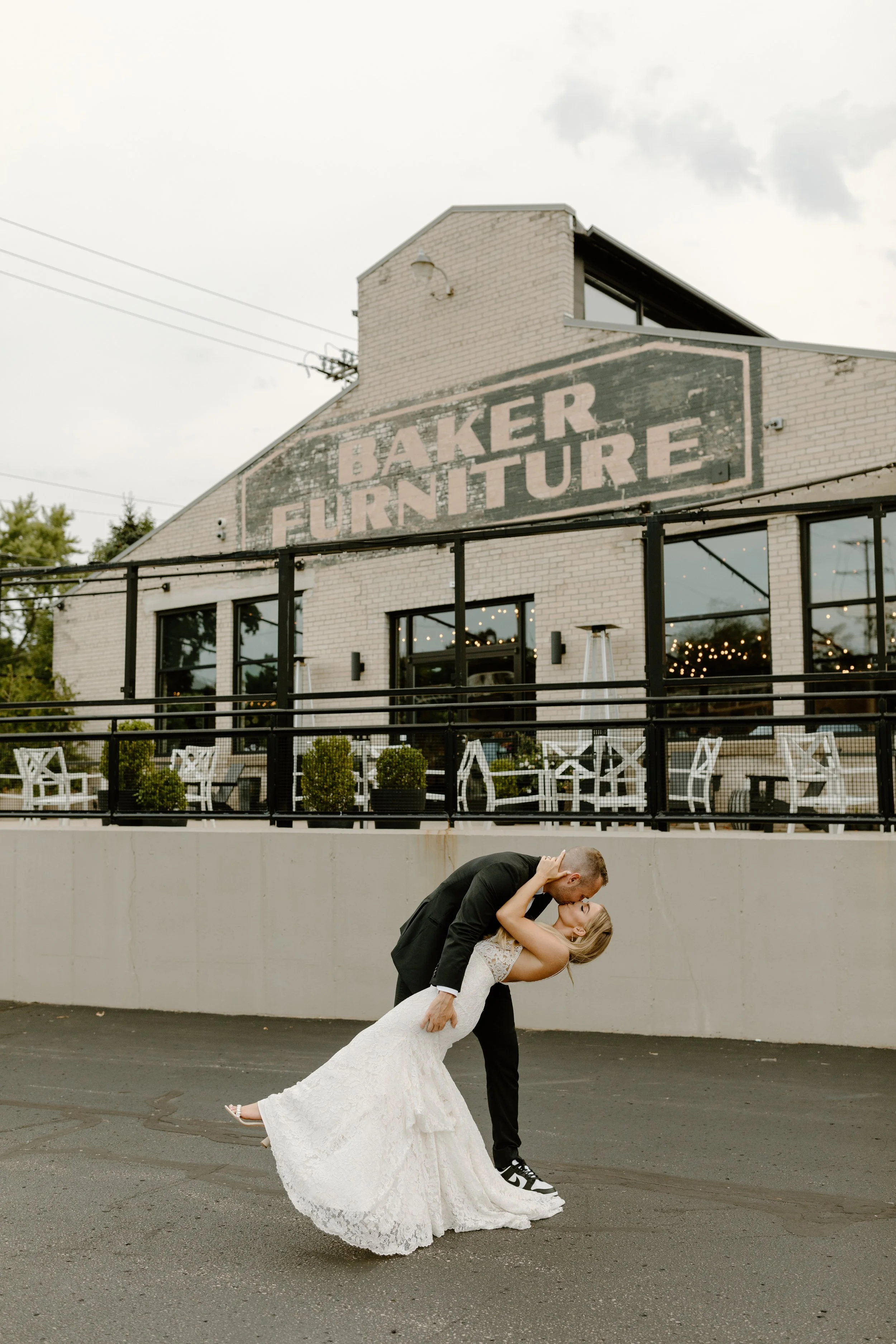 A couple dressed in wedding attire dancing in an outdoor urban area. The groom in a black suit and sneakers dips the bride in a white lace wedding dress in front of a white brick building with large windows and a sign that reads "Baker Furniture." Th